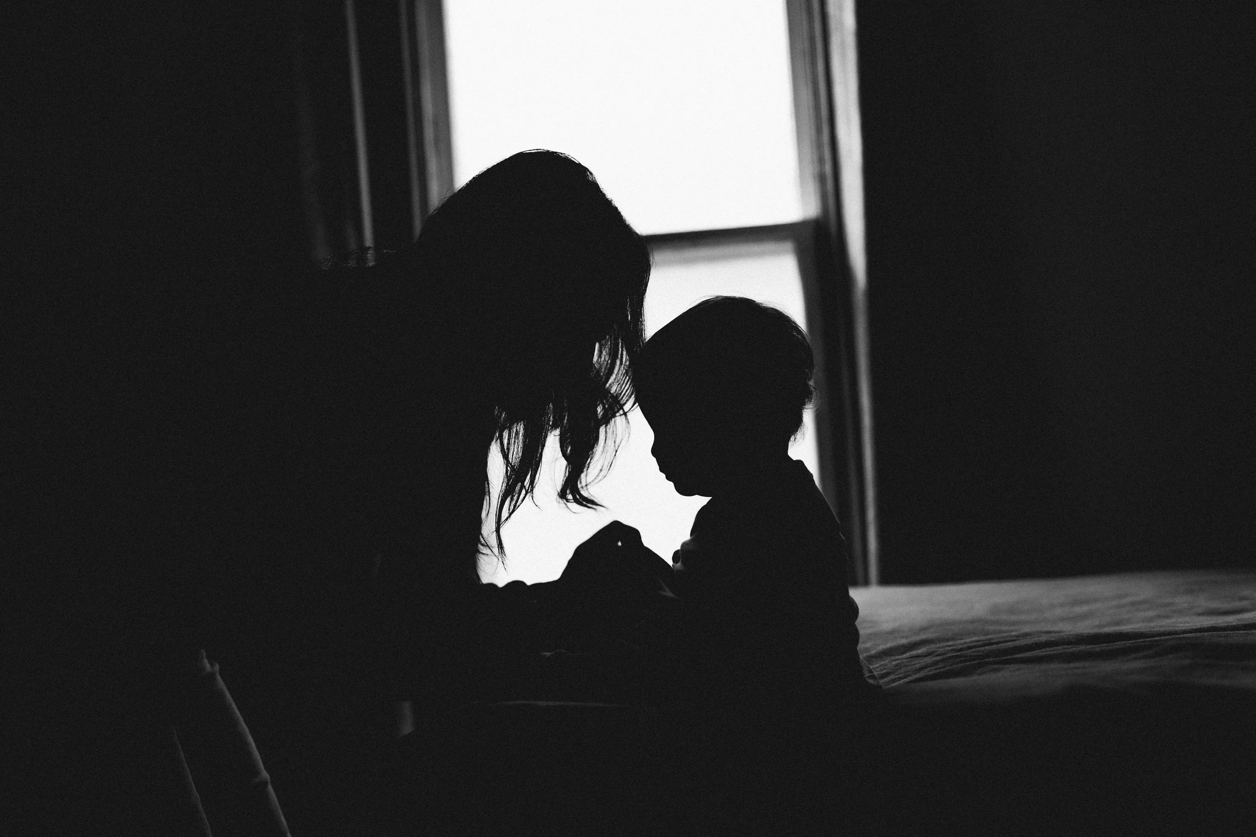 Silhouettes of a woman and a child sitting together near a window, touching foreheads in a tender moment.