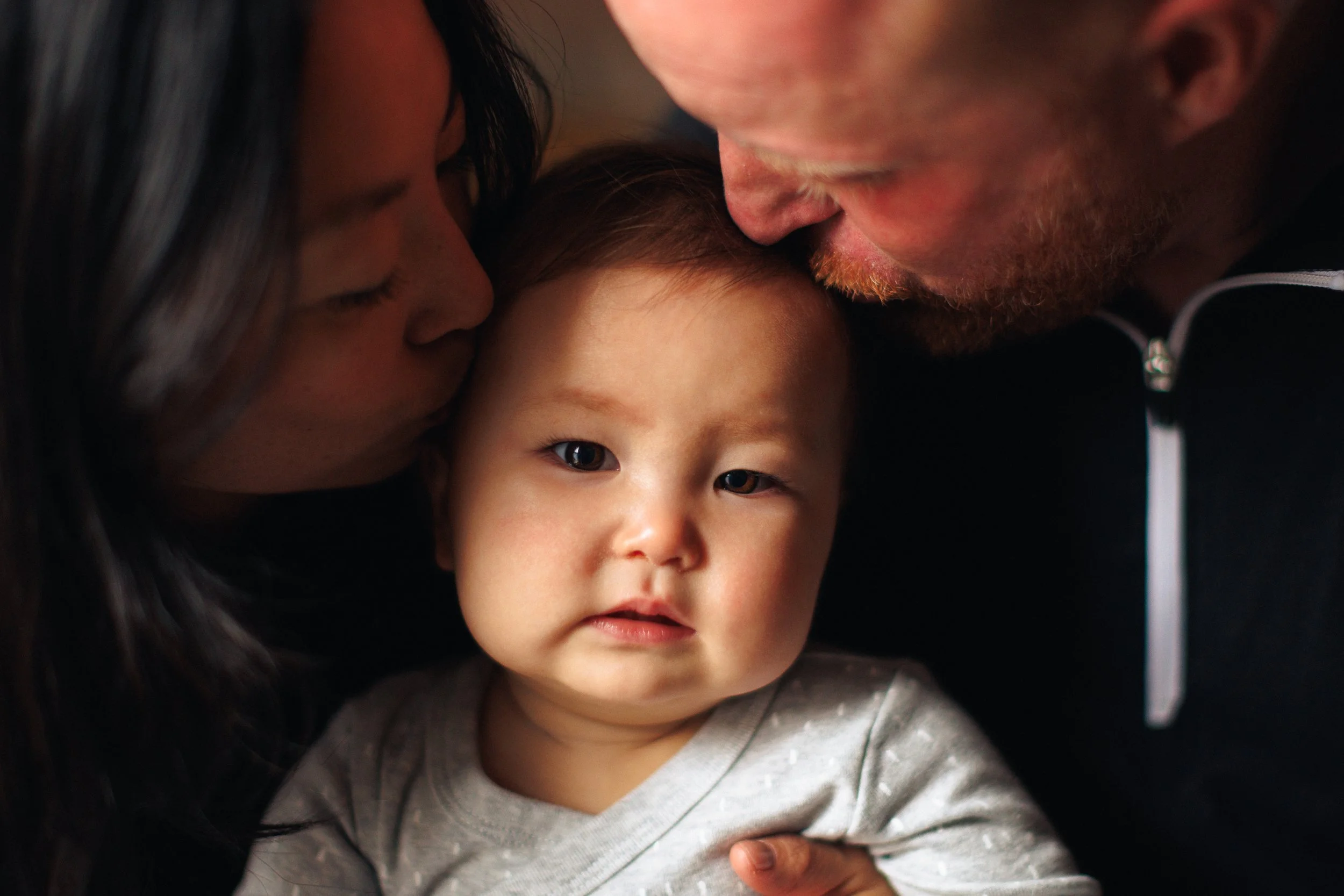 A young child with dark hair looking directly at the camera, being kissed on both cheeks by a woman and a man, who are likely the child's parents.