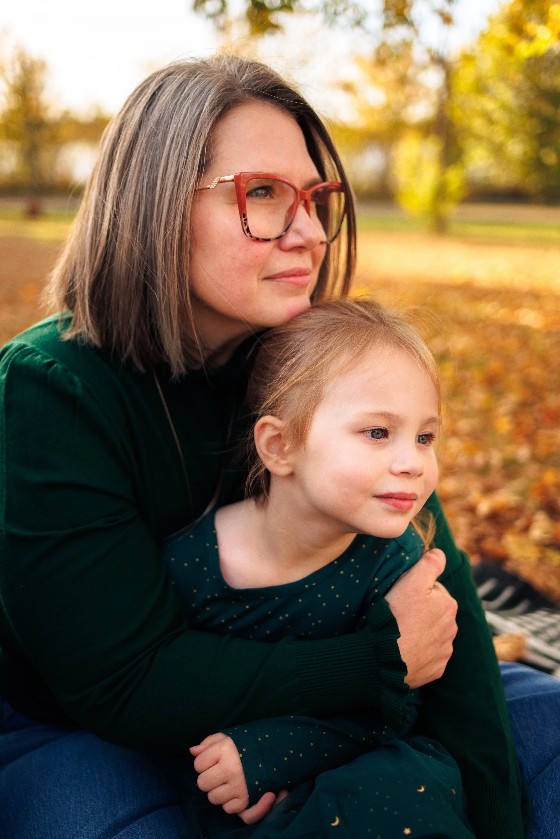 A woman with glasses and a young girl sitting outdoors in a park during autumn, surrounded by fallen leaves and trees with yellow and orange foliage.
