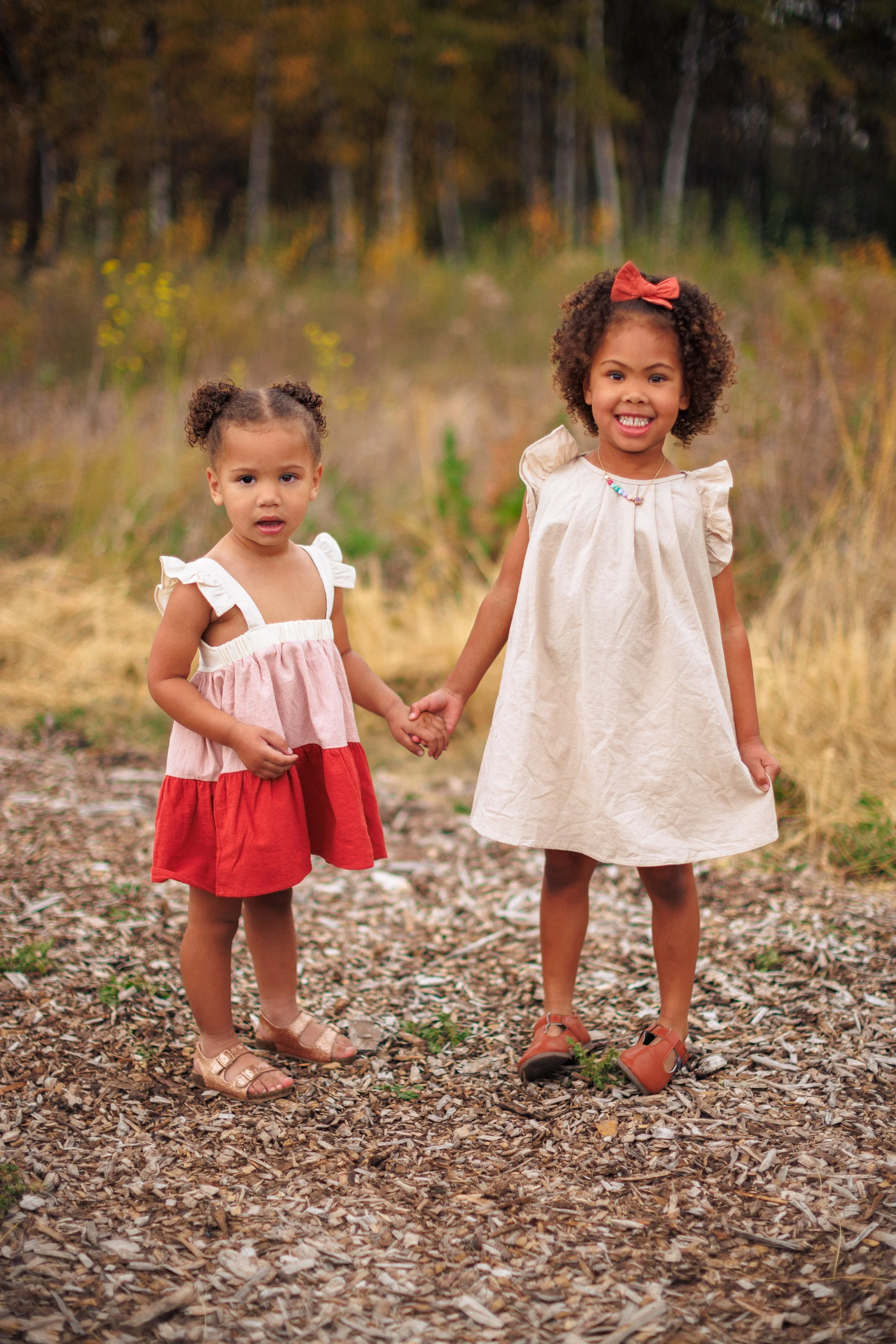 Two young girls holding hands outdoors on a trail with trees and yellow foliage in the background, one girl smiling and the other looking surprised.