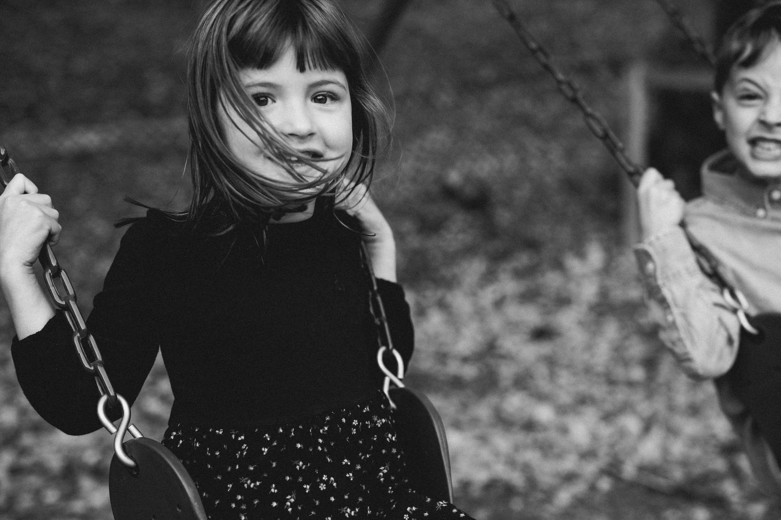 Black and white photo of a young girl and boy playing on swings outdoors, with the girl looking at the camera and the boy smiling in the background.