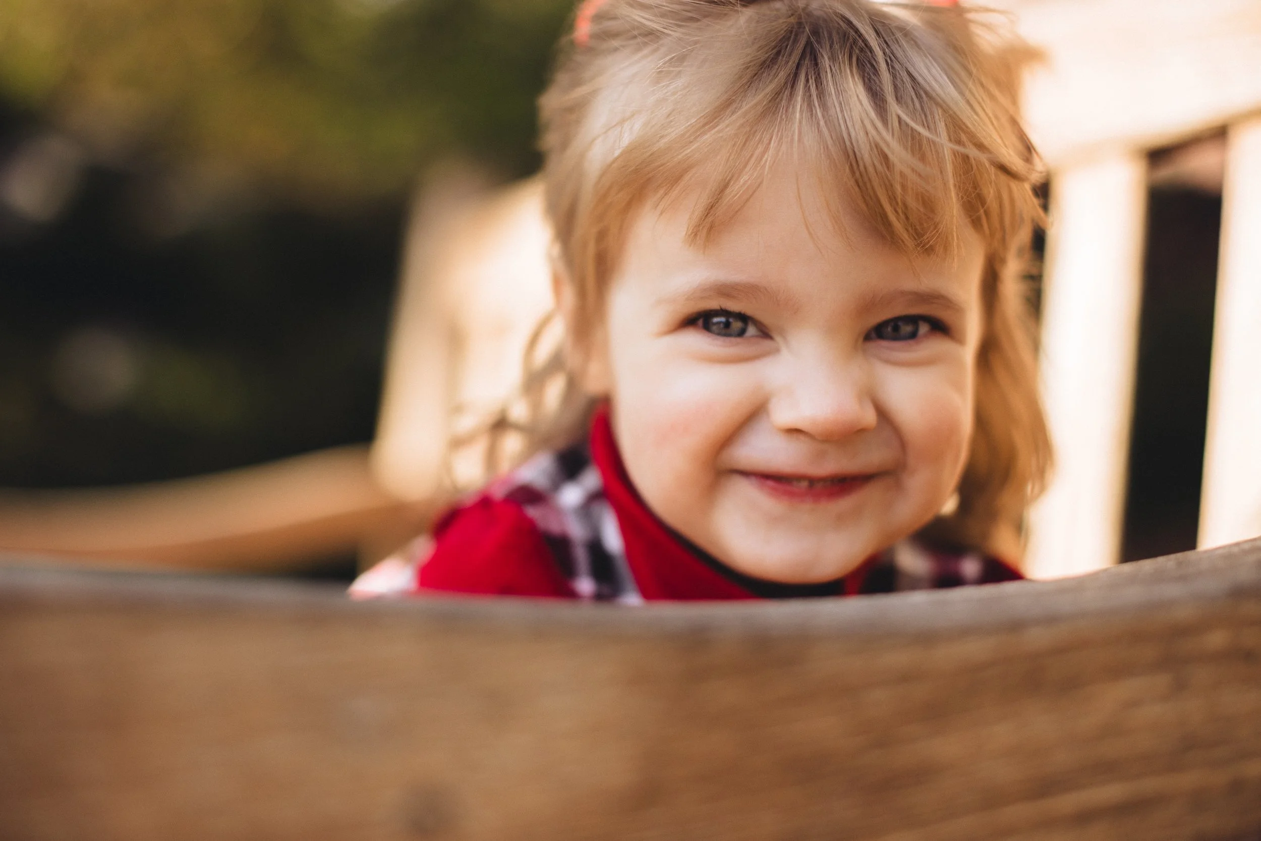 Close-up of smiling young girl with light brown hair, wearing a red shirt and plaid collar, outdoors with blurred background.