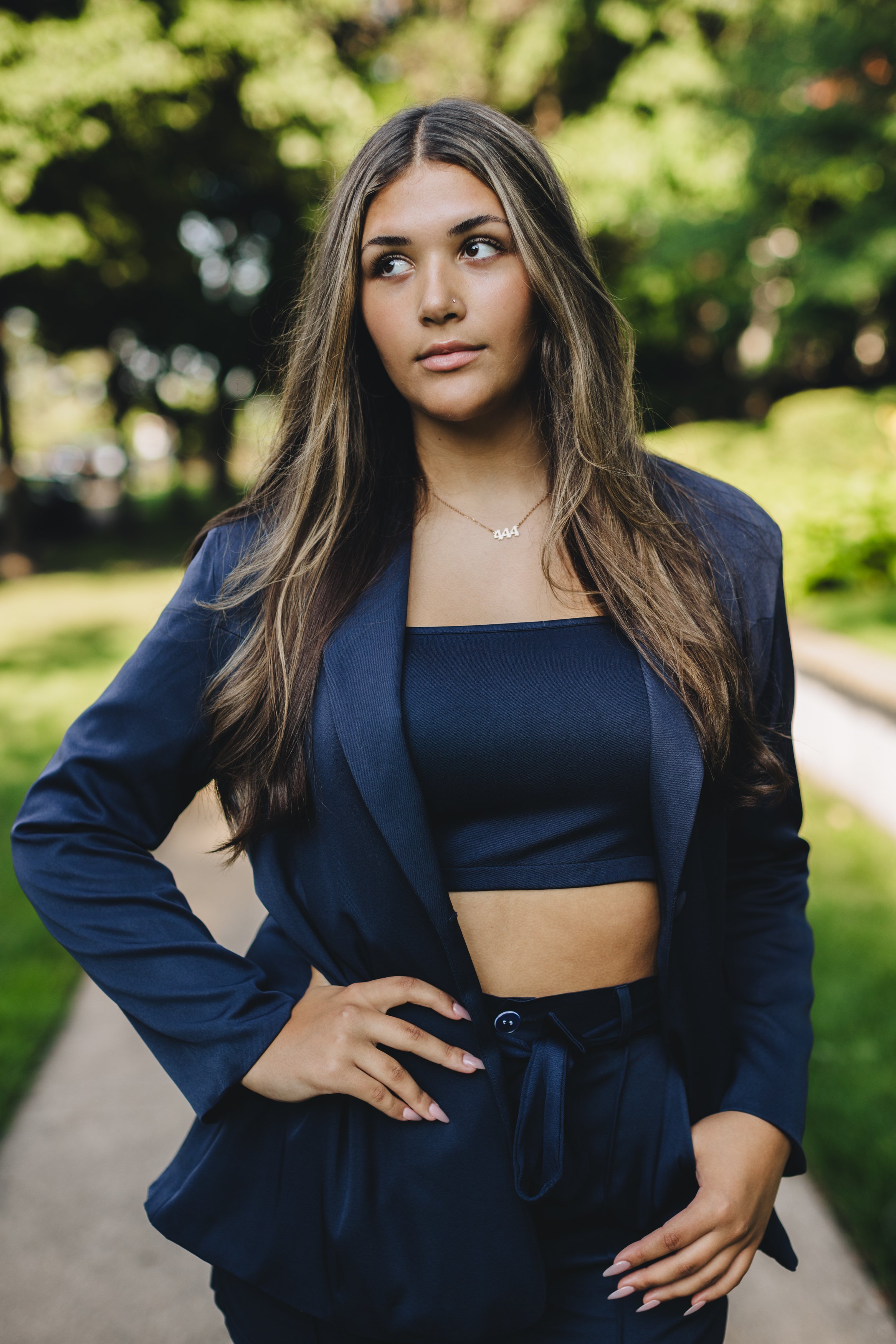 A young woman with long brown hair standing outdoors on a sidewalk, wearing a navy blazer and matching cropped top, with trees and greenery in the background.
