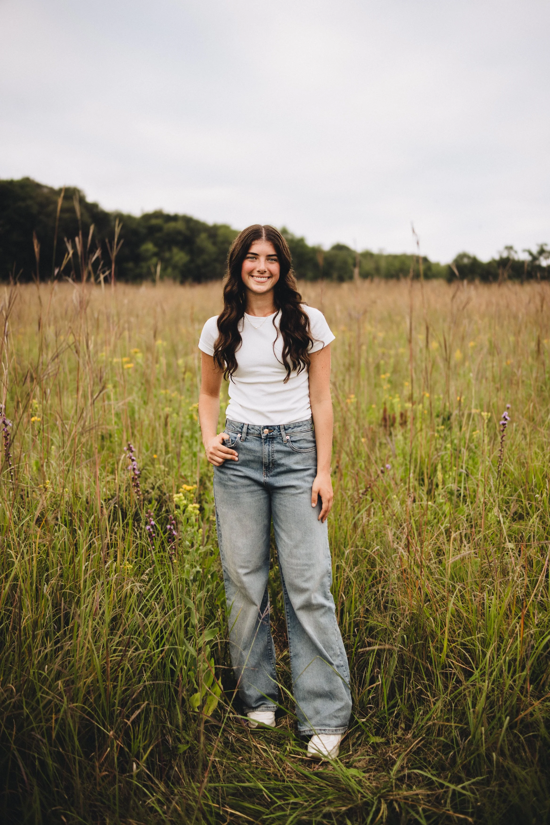 A young woman with long dark hair, wearing a white t-shirt and baggy jeans, standing in a grassy field during daytime, smiling at the camera.