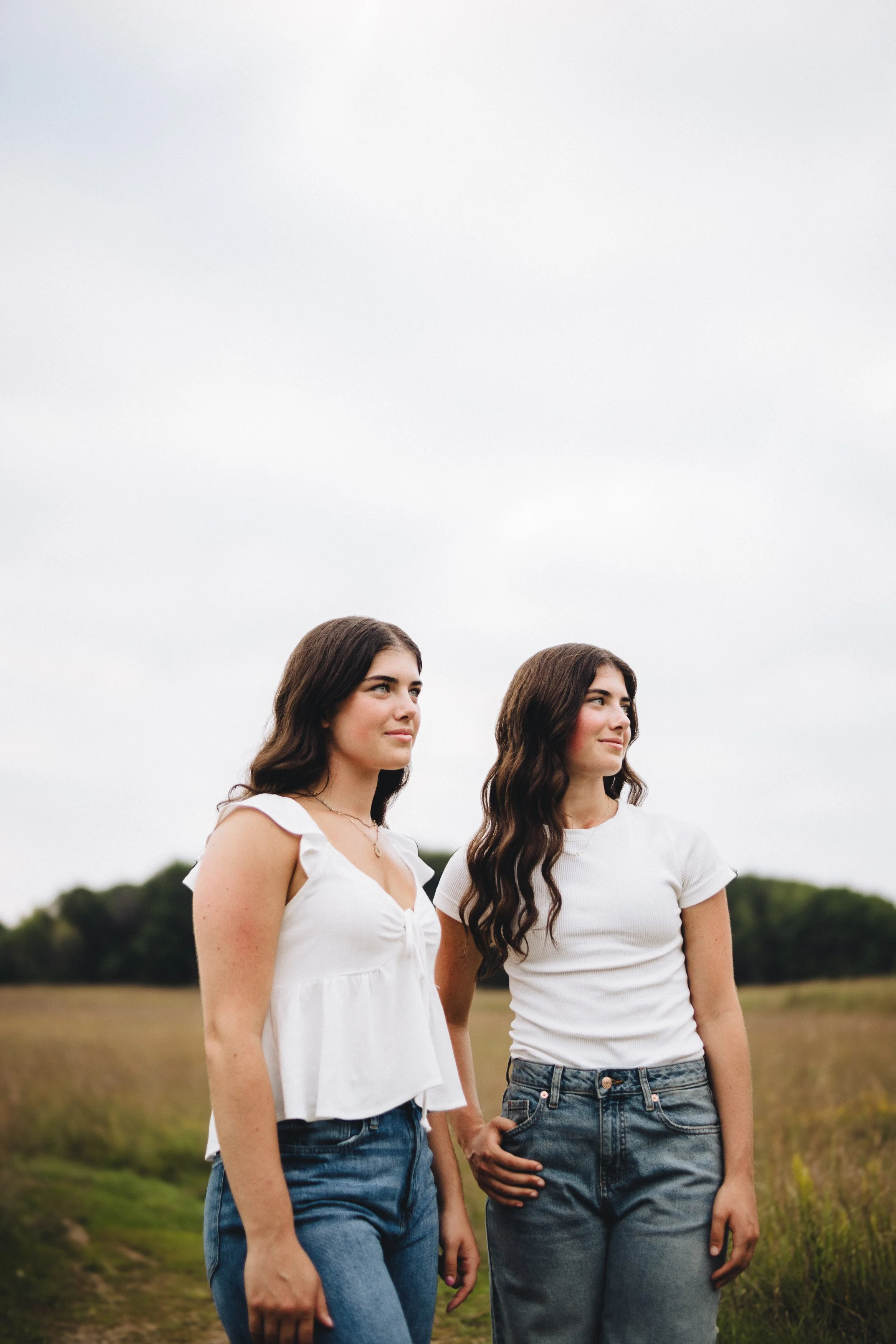 Two young women with long dark hair standing outdoors in a field with cloudy sky, wearing casual white tops and jeans.