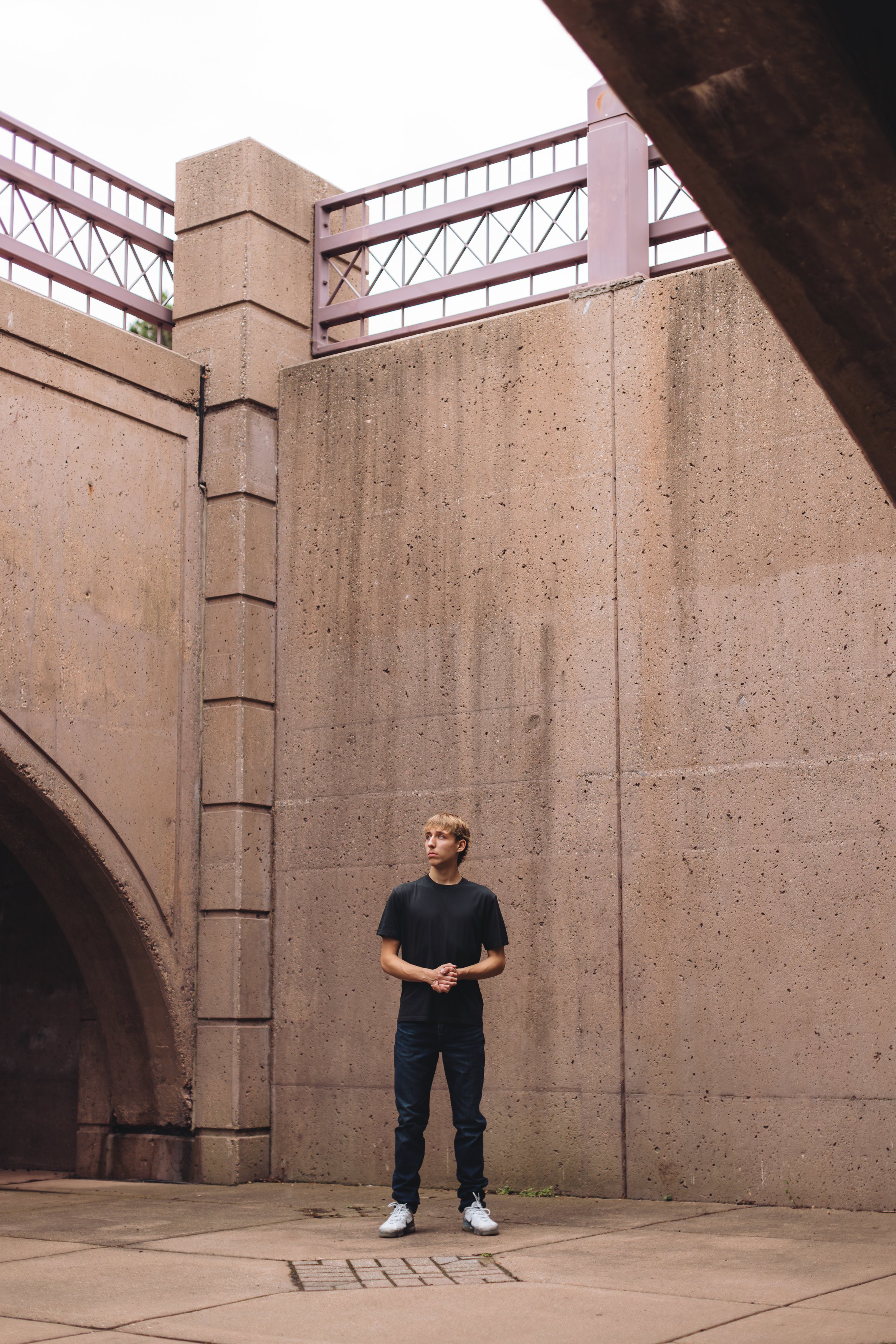 A young man with blonde hair, wearing a black t-shirt, dark jeans, and white sneakers, stands alone outdoors against a large beige concrete wall. The sky is overcast, and part of an archway and a railing are visible in the background.