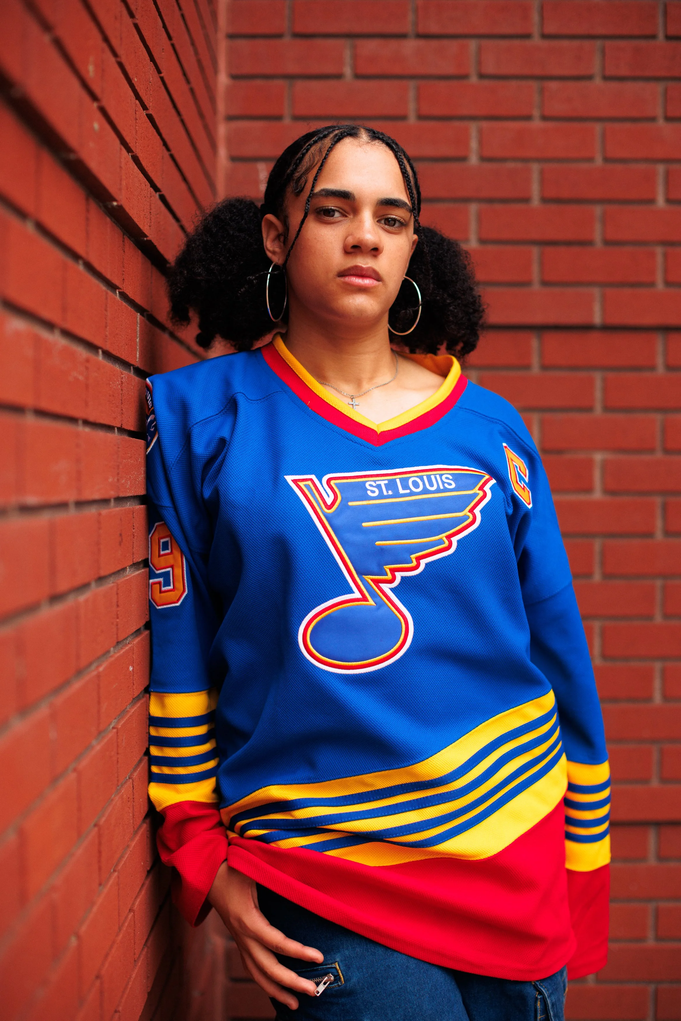 A young woman with curly hair in pigtails wearing a blue St. Louis hockey jersey, standing against a brick wall.