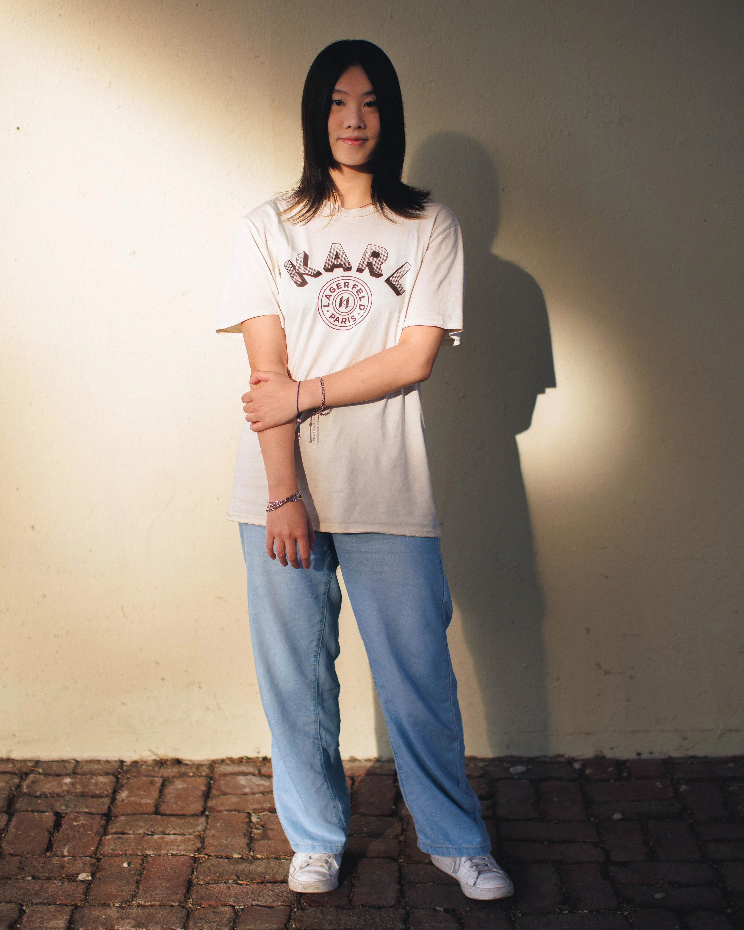 A young woman with shoulder-length black hair standing against a beige wall, wearing a white T-shirt with the word "KARL" and a circular logo, light blue jeans, and white sneakers, with her arms crossed and casting a shadow on the wall.