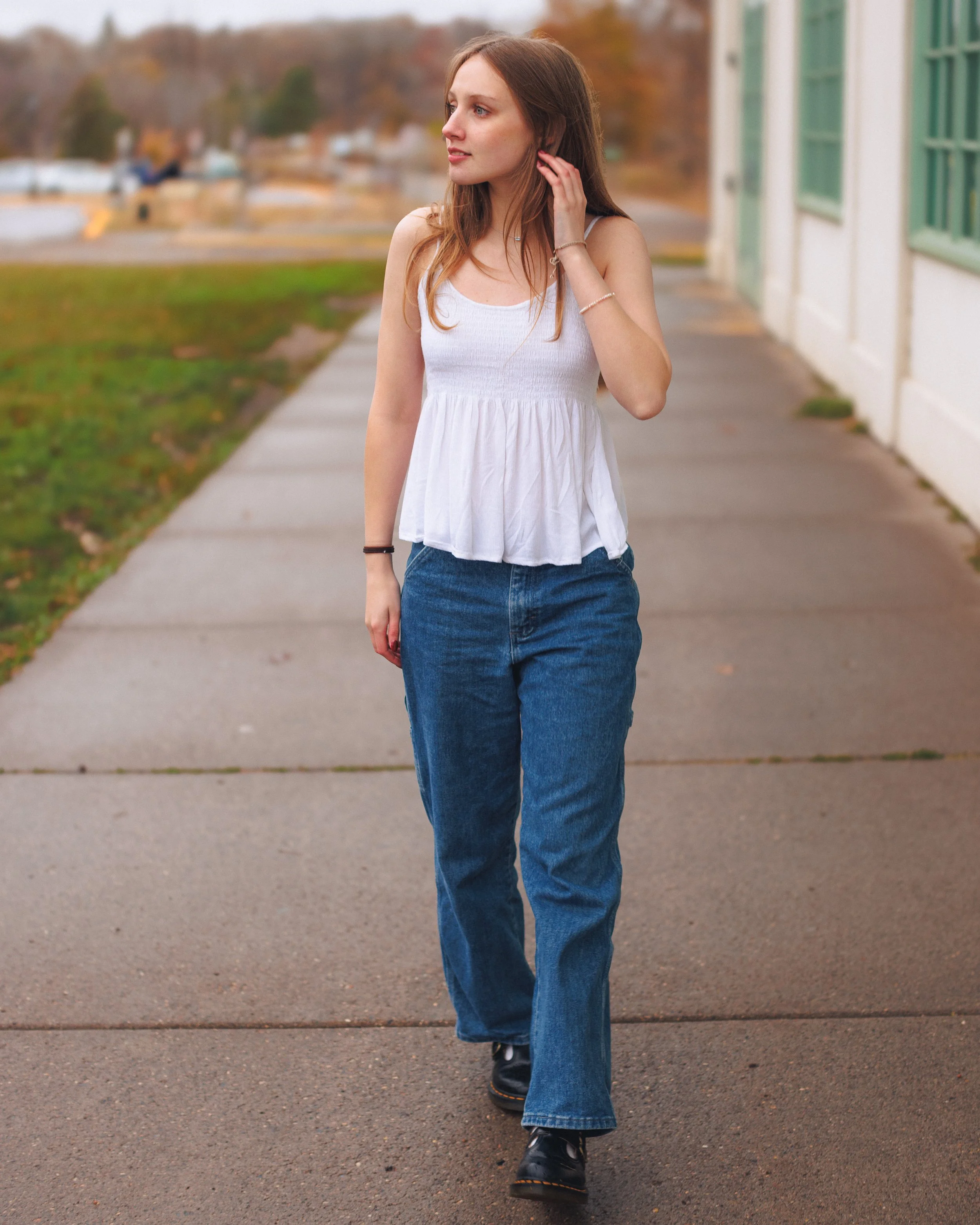 A young woman walking on a sidewalk during autumn, wearing a white sleeveless top, blue jeans, and black shoes, with fall foliage in the background.