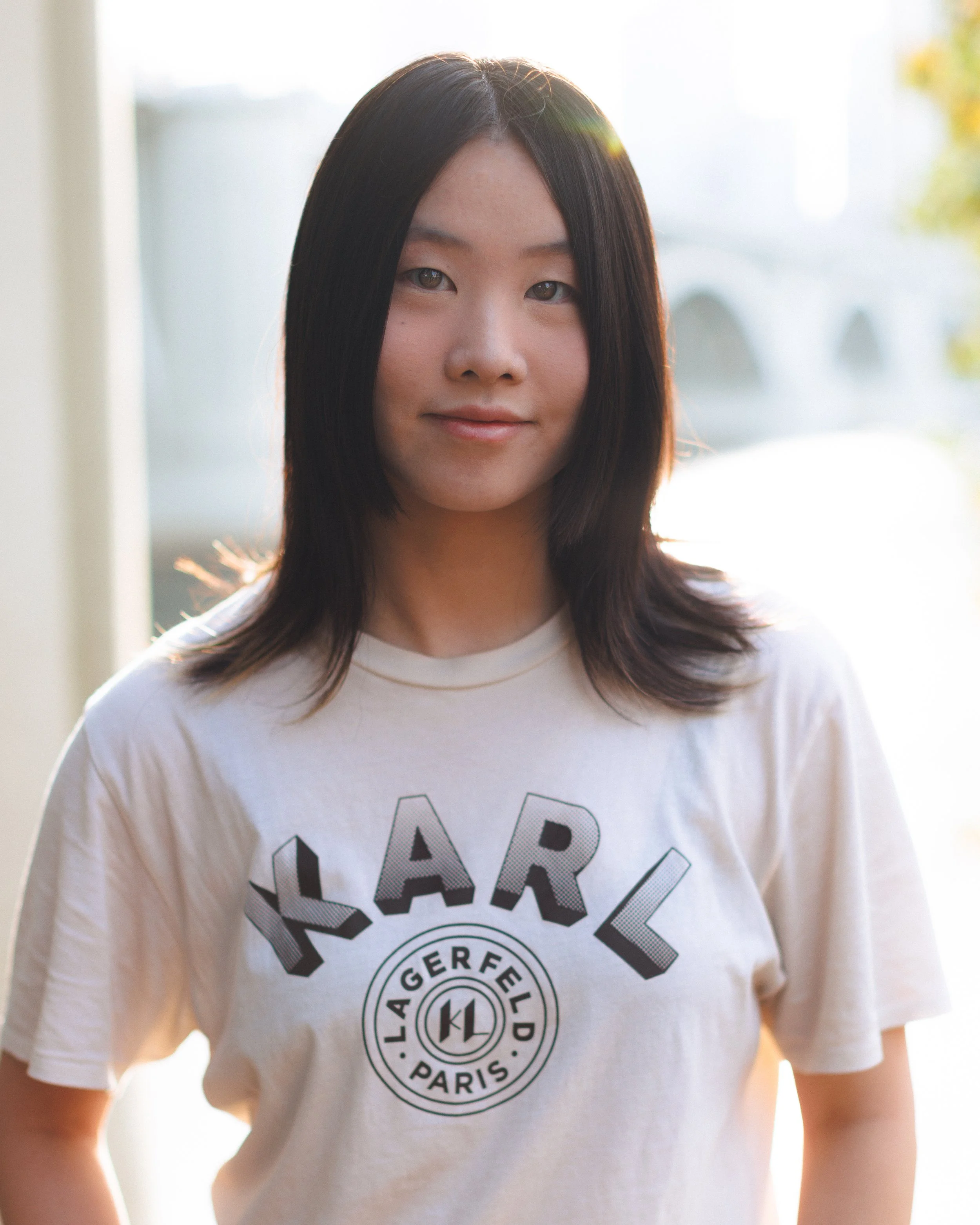 Young woman standing indoors near a window, wearing a white T-shirt with the word 'LARL' and a circular logo with the words 'LAGERFELD PARIS' on it, illuminated by natural sunlight.