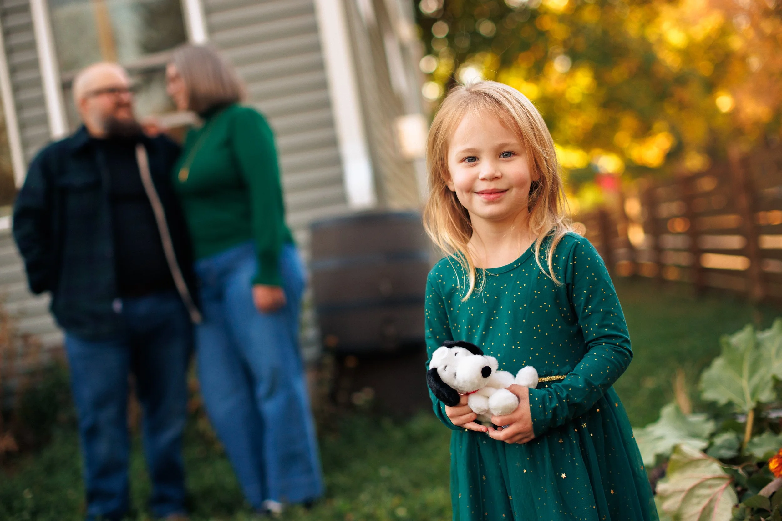 A smiling young girl with blonde hair in a green dress holding a Snoopy stuffed animal, outdoors with a blurred background of a house, a fence, and two adults talking in the background.