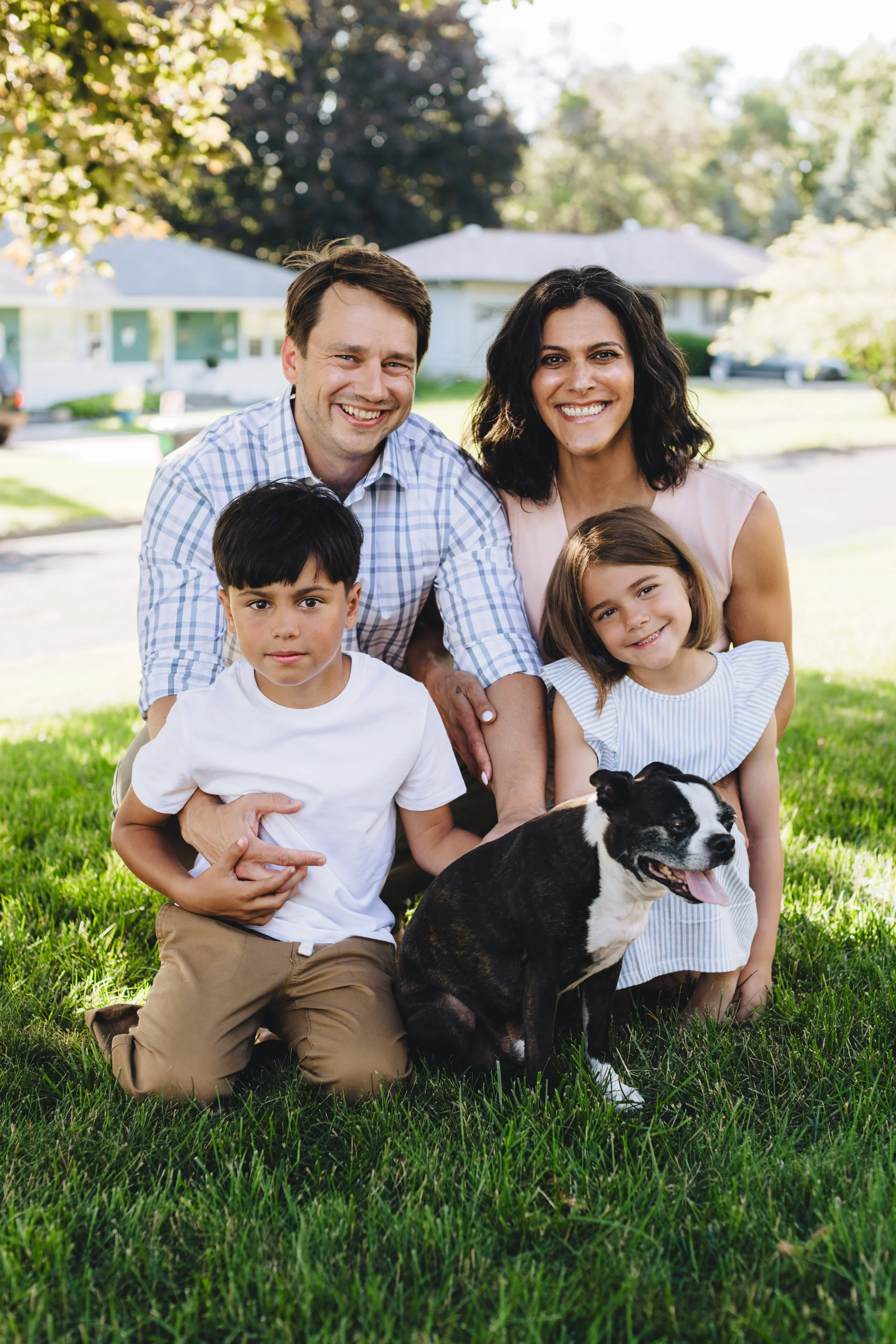 A happy family of four with a dog outdoors in a park on a sunny day. The family includes a man, woman, young boy, and girl, all smiling and smiling with trees and houses in the background.