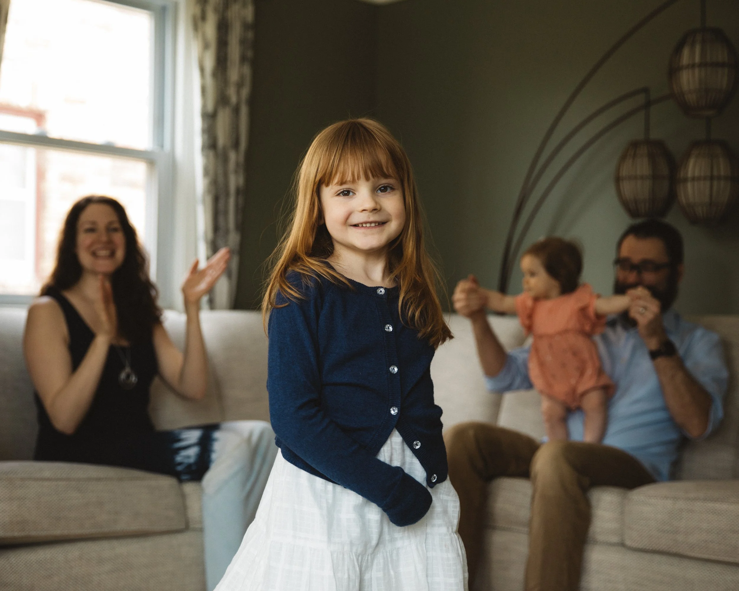 A young girl with red hair smiling in front of her family in a living room, with a woman and a man playing with a toddler in the background.