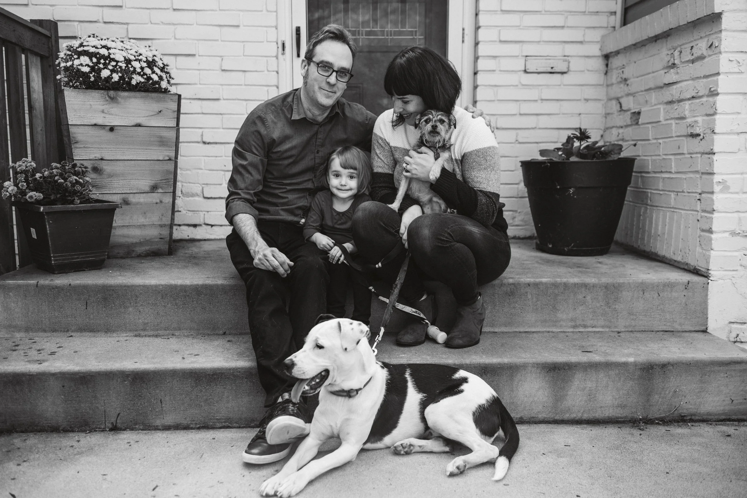 A black and white photo of a family sitting on the front steps of their house, with a man, woman, young girl, two dogs, and potted plants around them.