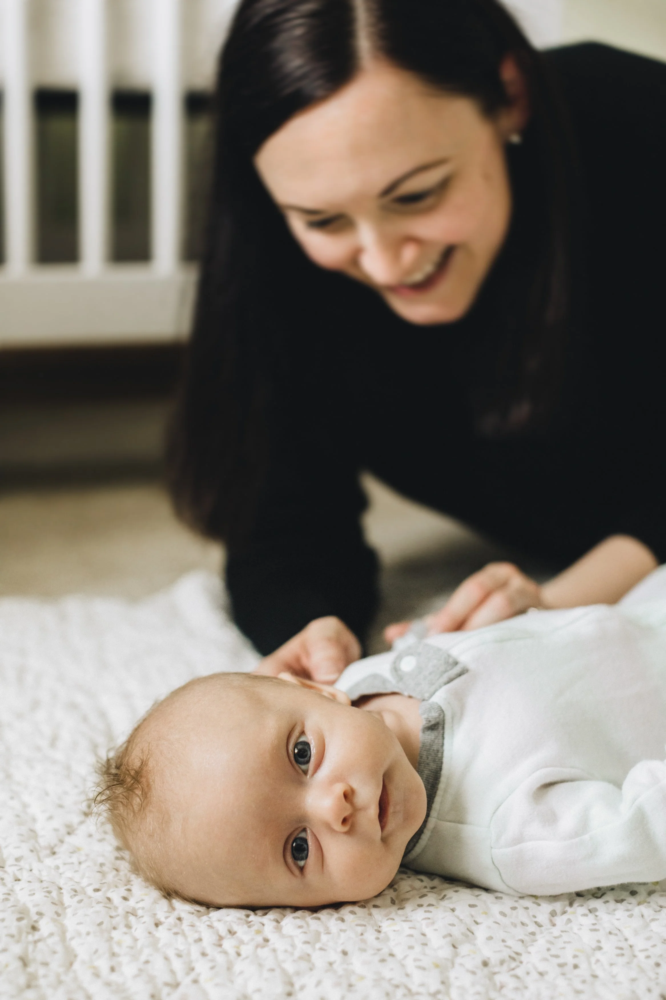 A woman with dark hair smiles joyfully while laying on a cream-colored blanket, looking at a baby who is lying on the blanket, gazing towards the camera.