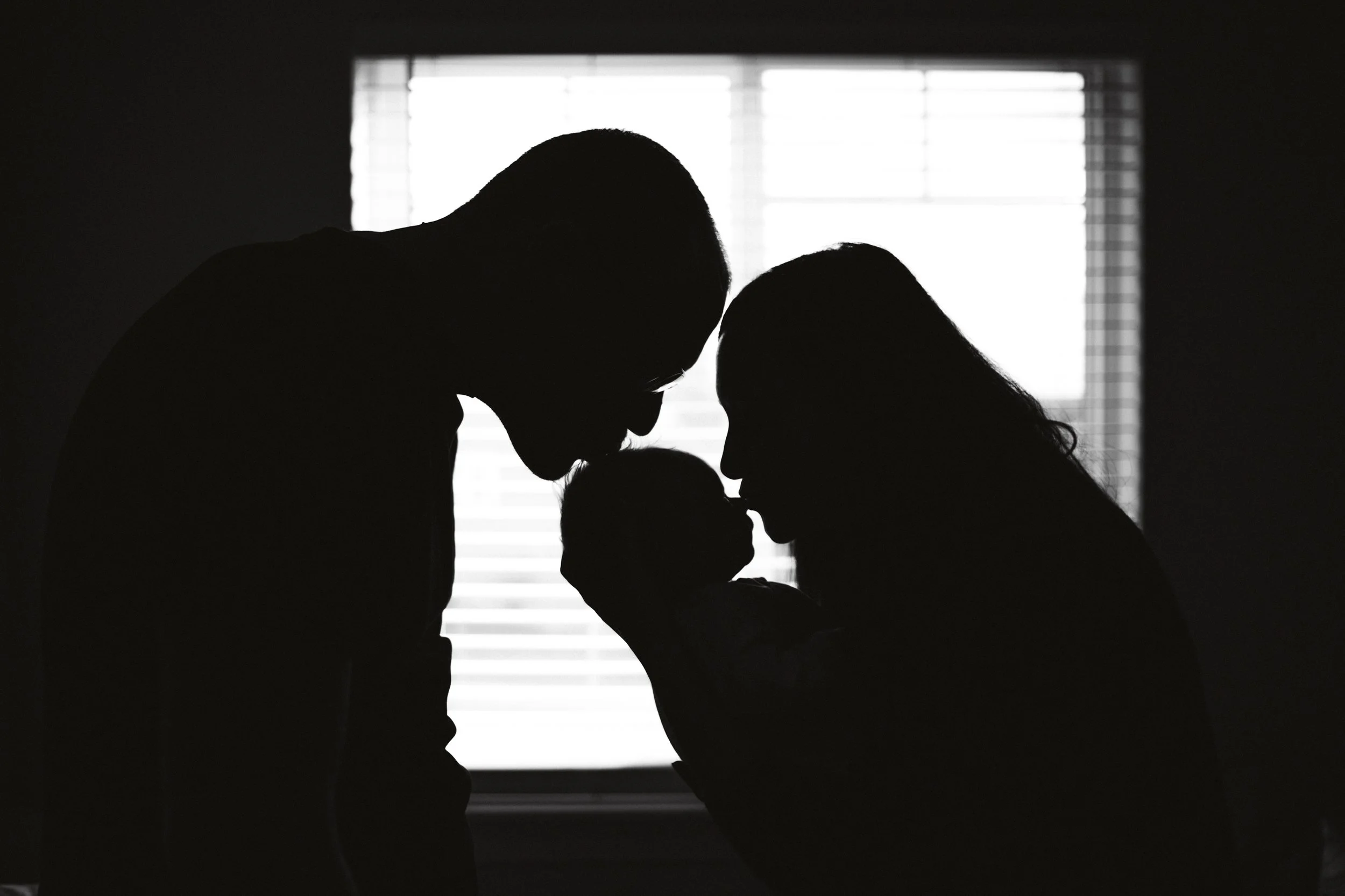 Silhouette of a family with a baby, with faces close together, backlit by a window.