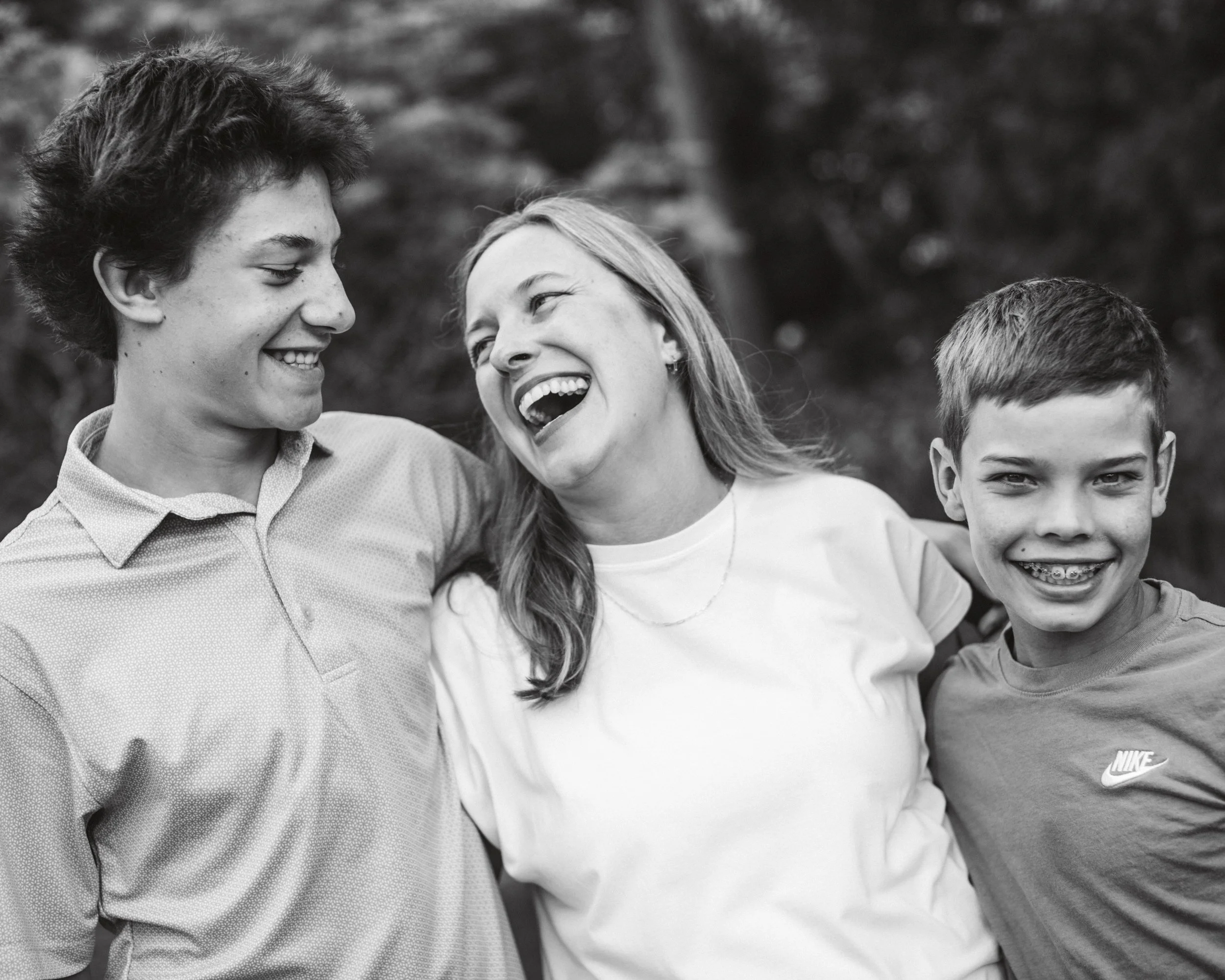 Three people, two children and a woman, are outside, smiling and laughing together, with their arms around each other.