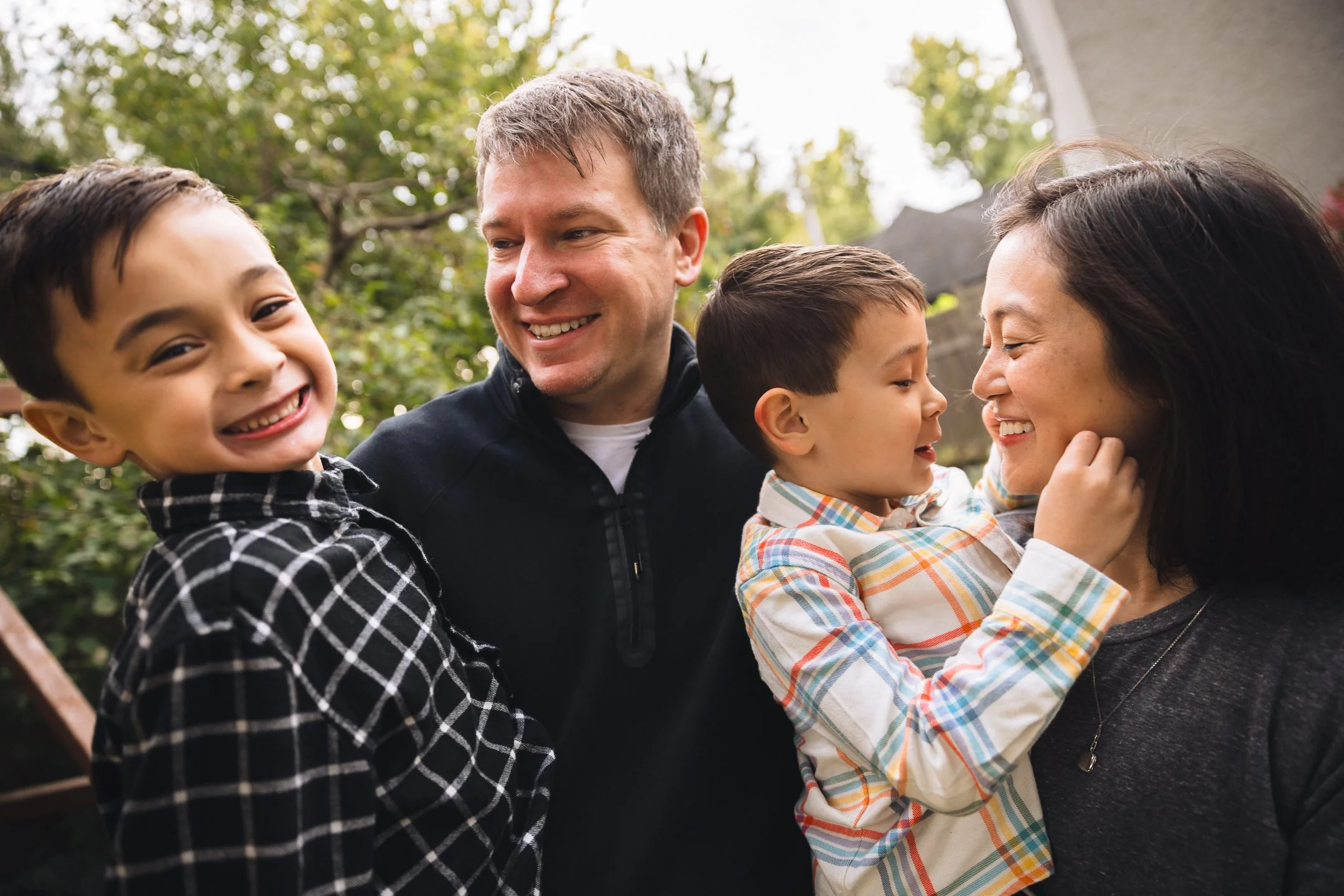 A family of four outside, smiling and interacting, with a woman and two young boys. The woman and one of the boys are touching faces, and all appear happy.