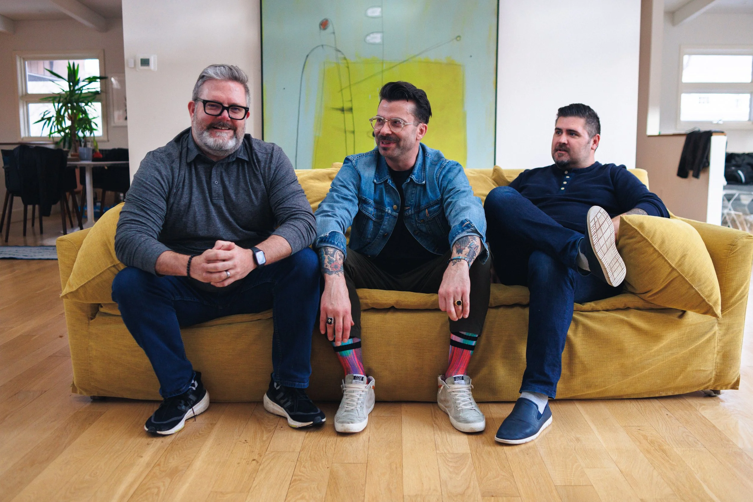 Three men sitting on a yellow sofa in a living room. They are casually dressed and appear to be having a conversation.