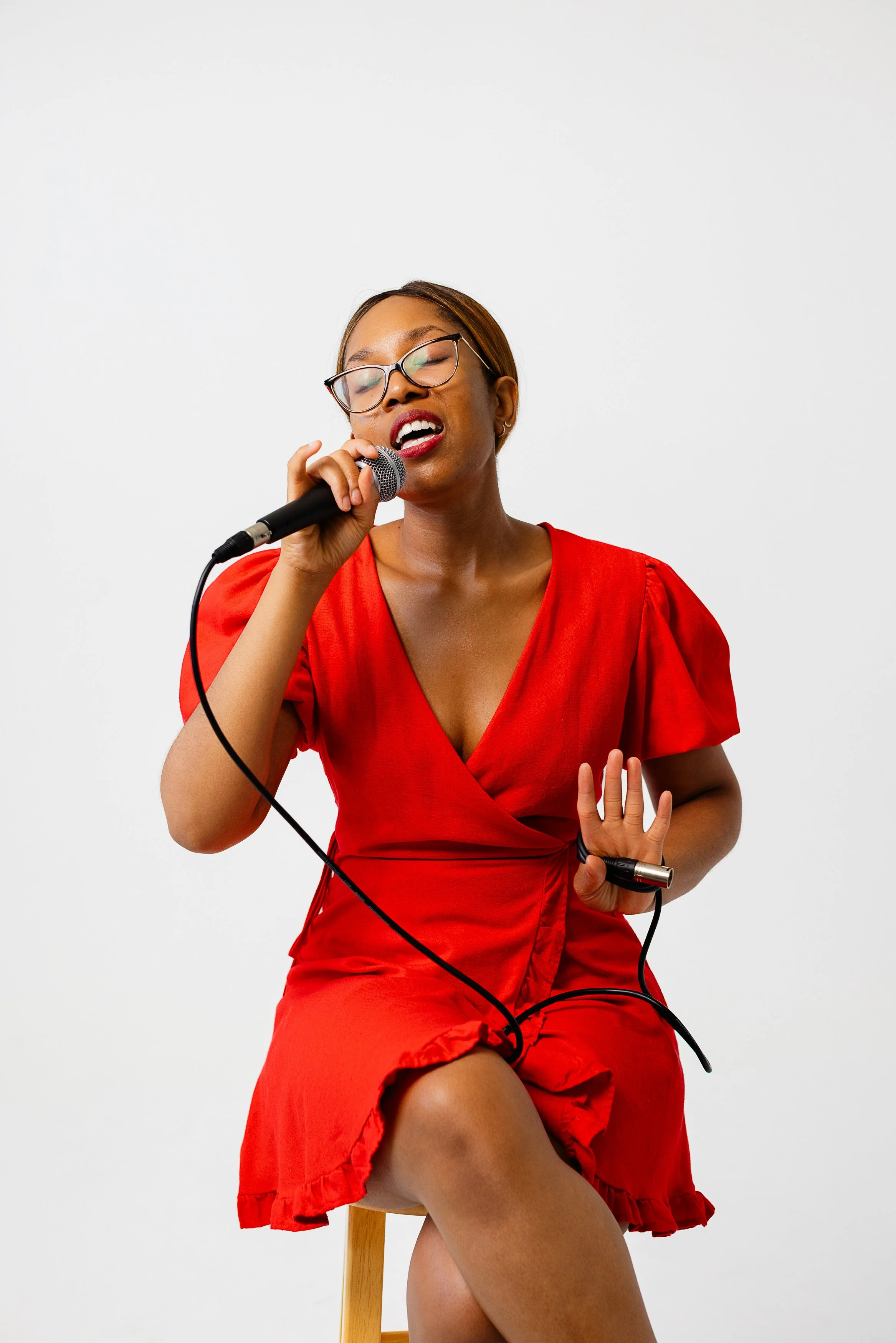 Woman in red dress singing into a microphone while sitting on a stool against a white background.