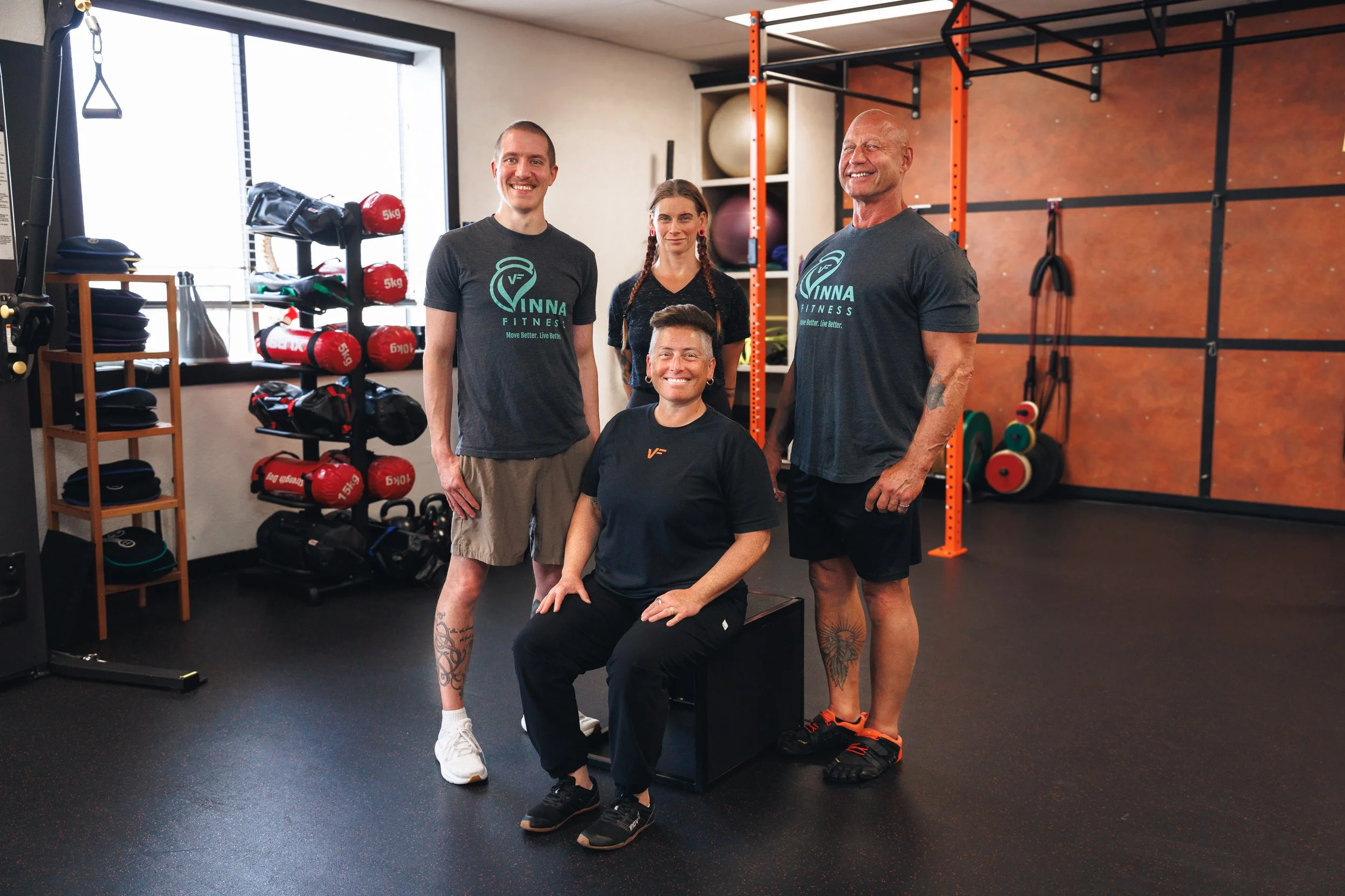 Group of four diverse individuals smiling in a gym, with gym equipment like weights and resistance bands in the background.