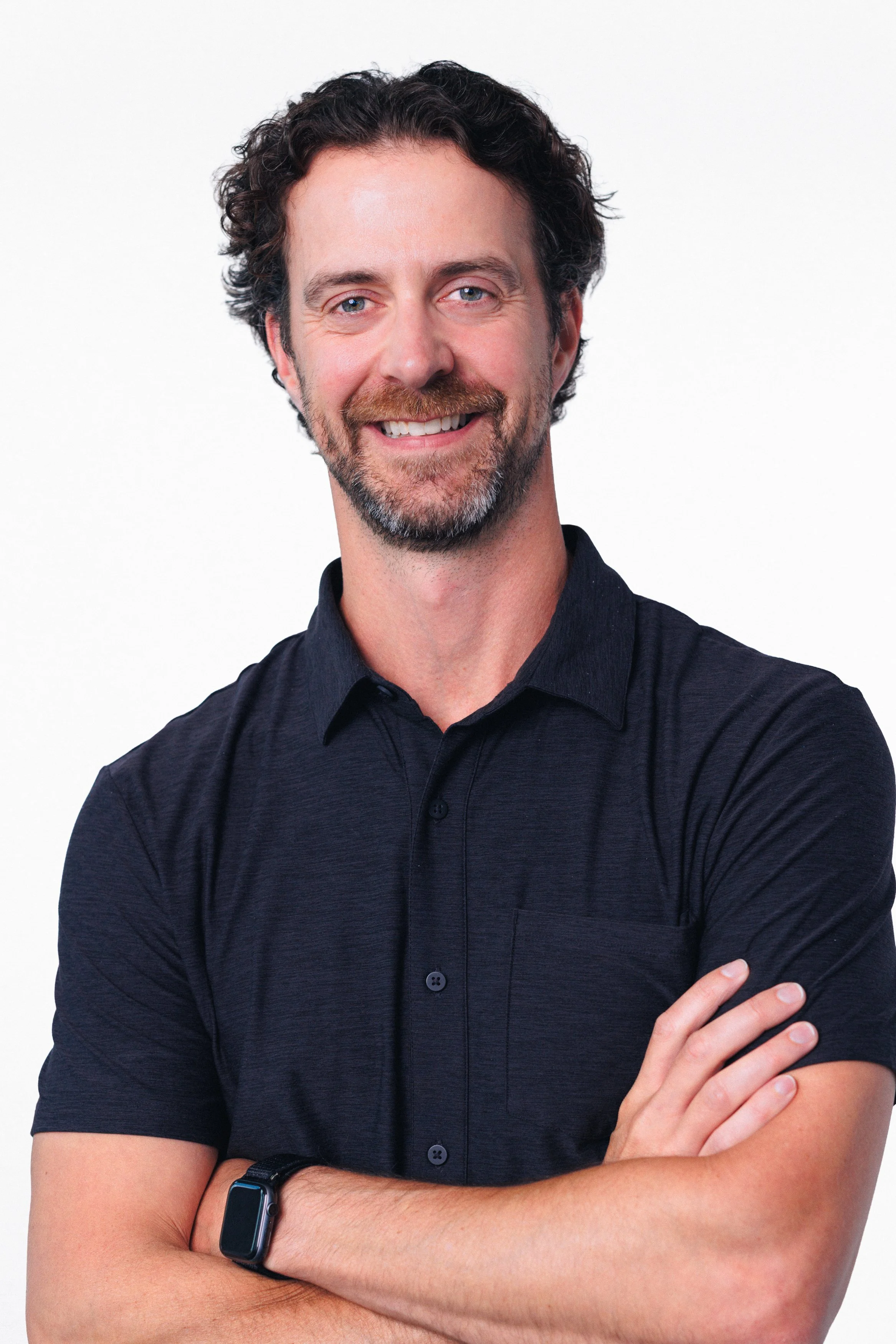 A man with curly dark hair and a beard, smiling, wearing a black shirt, with crossed arms and a smartwatch on his left wrist, standing against a white background.