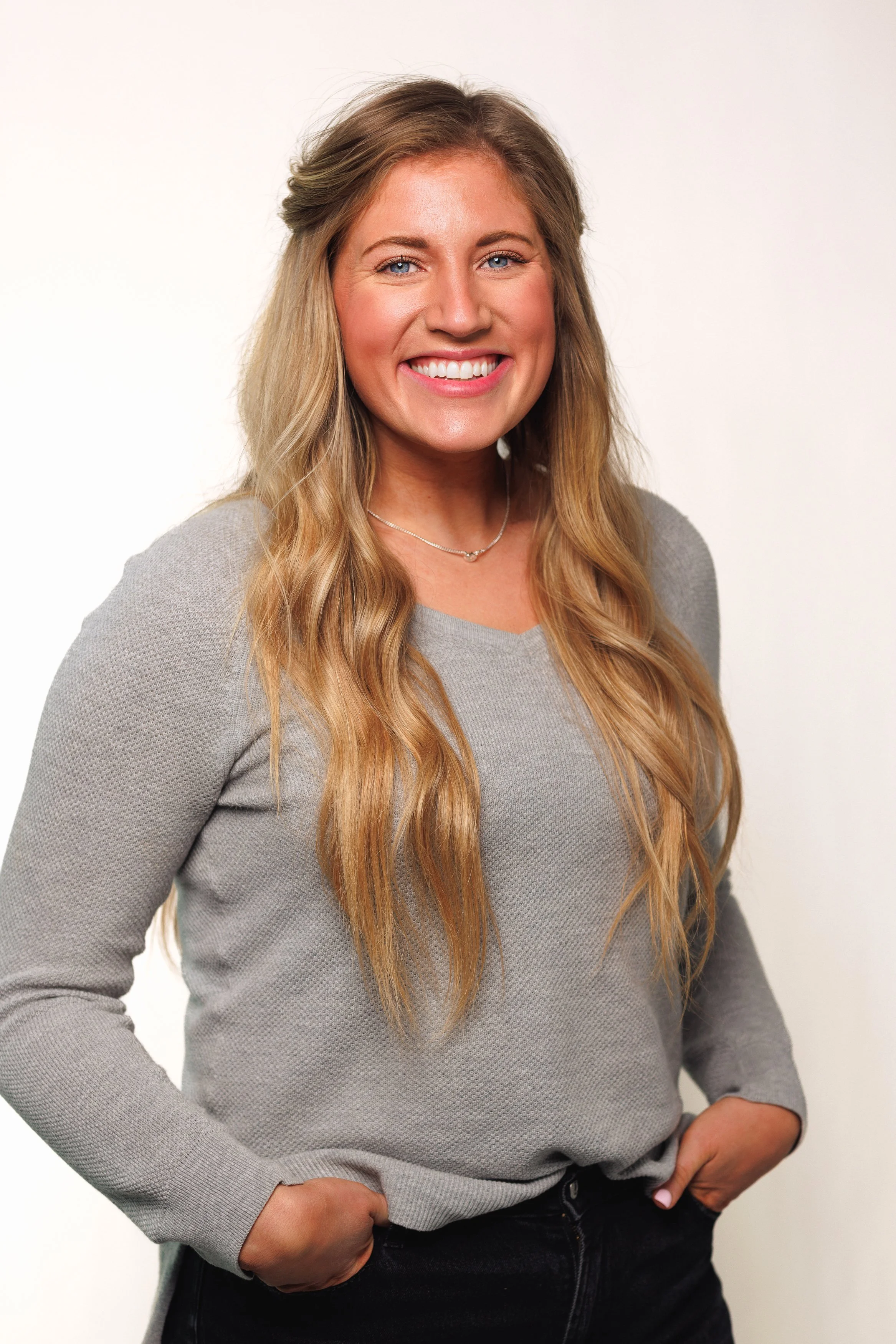 A woman with long wavy blonde hair, smiling and wearing a light gray long-sleeve sweater and a necklace, stands against a plain white background.