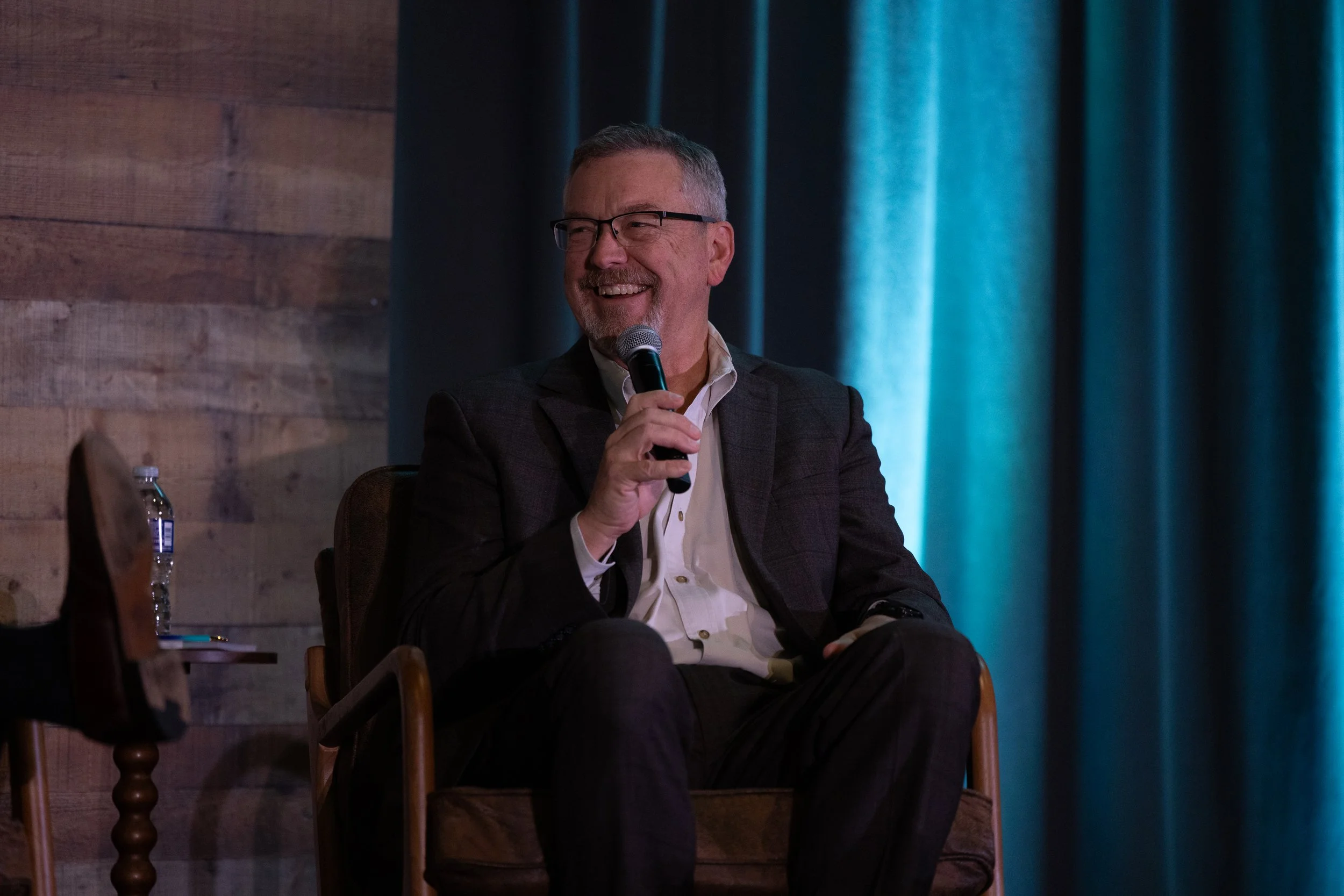 A middle-aged man with glasses, wearing a suit and open-collared shirt, sitting on a wooden chair, smiling, and holding a microphone during a presentation or discussion. There is a water bottle on a small table beside him, with dark curtains and a wo