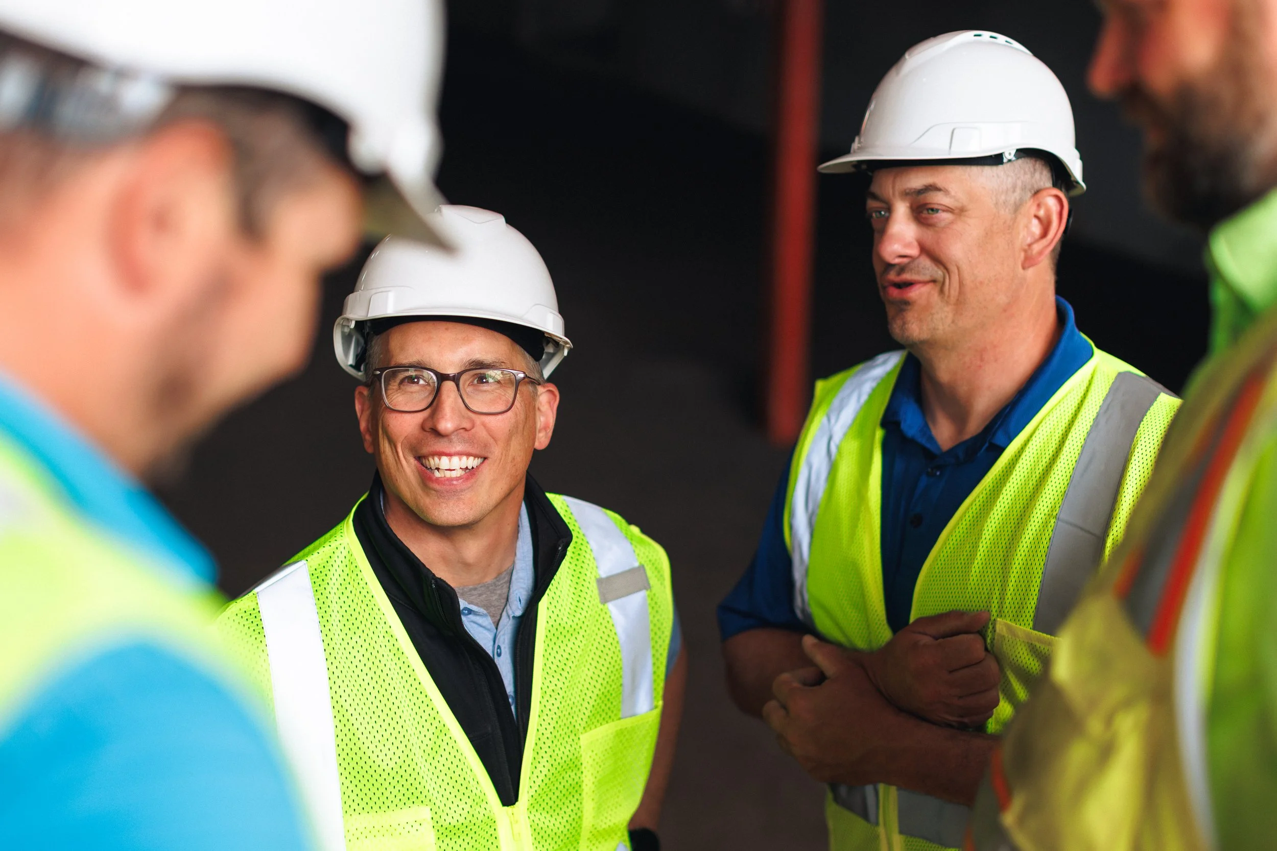 Group of construction workers wearing white hard hats and high-visibility vests, engaged in conversation on a construction site.