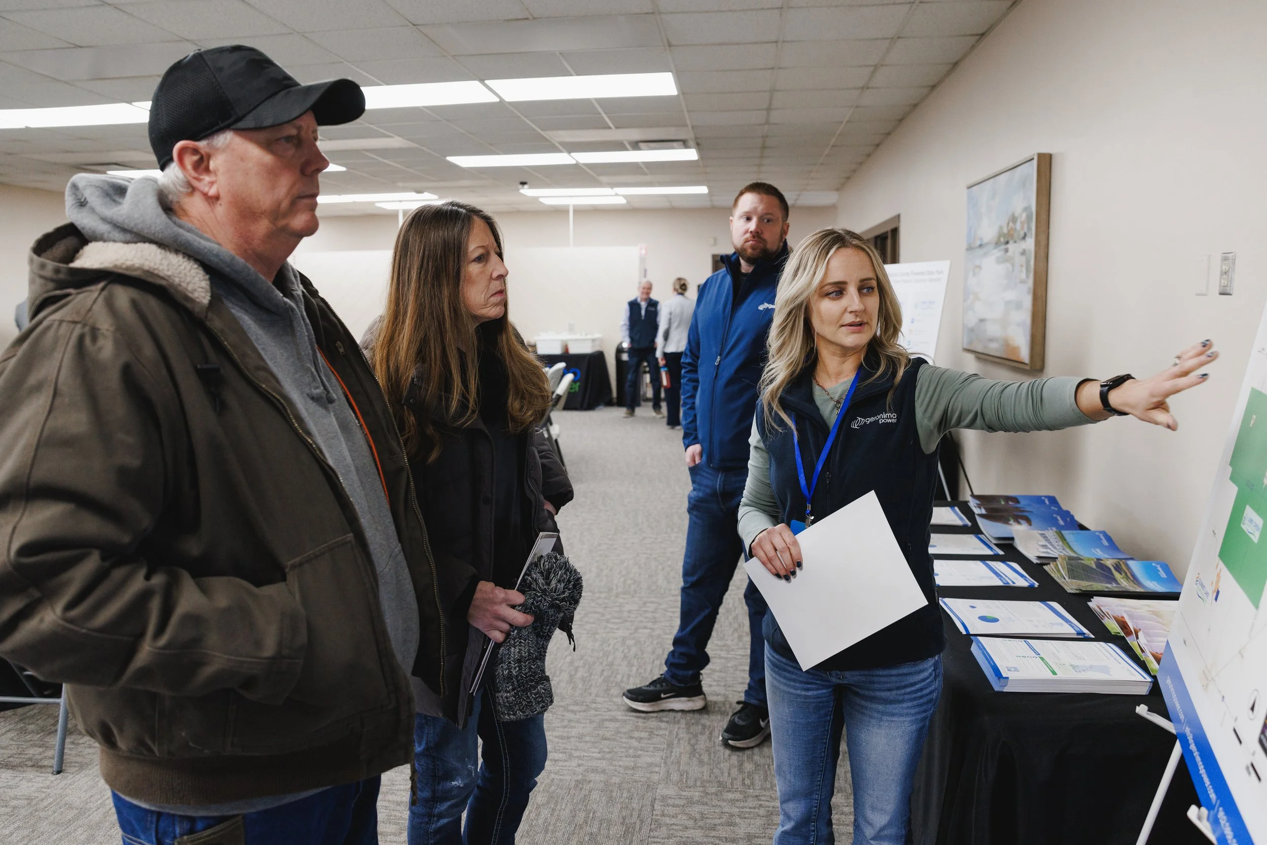 A woman presenter is explaining a display board to three attendees at an indoor event or conference, with additional attendees and display tables visible in the background.