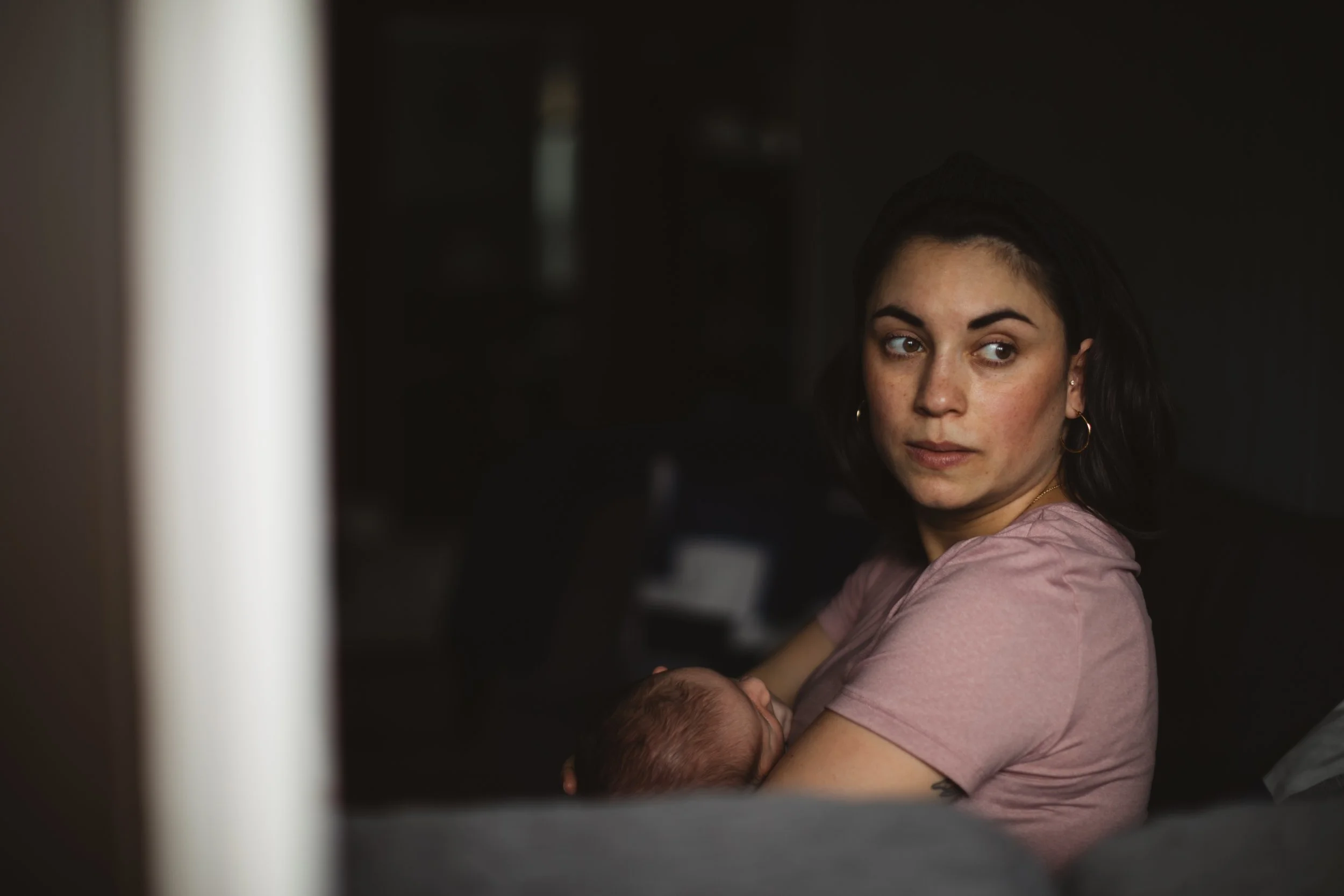 A woman with dark hair, wearing a pink shirt, holding a sleeping baby in her arms, sitting on a dark couch in a dimly lit room.