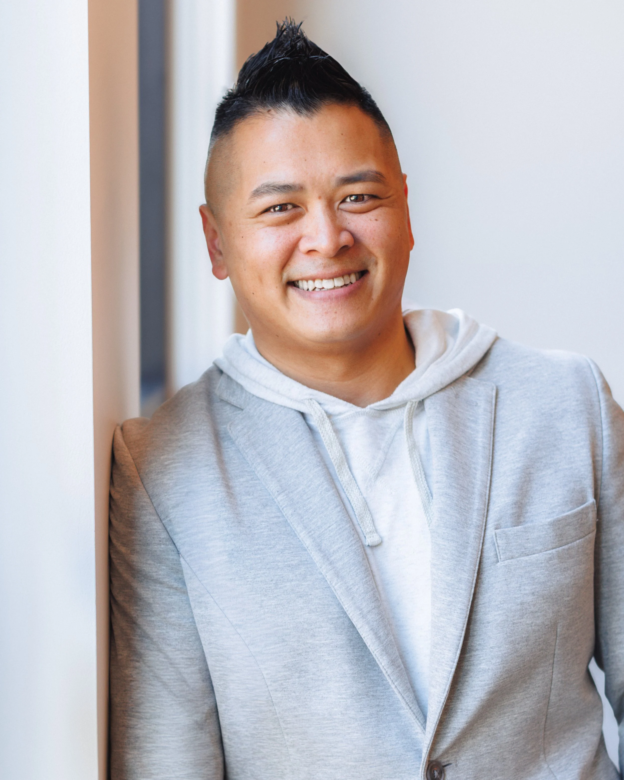 A smiling man with short dark hair styled upwards, wearing a light gray hoodie, leaning against a white wall near window, with soft natural light.