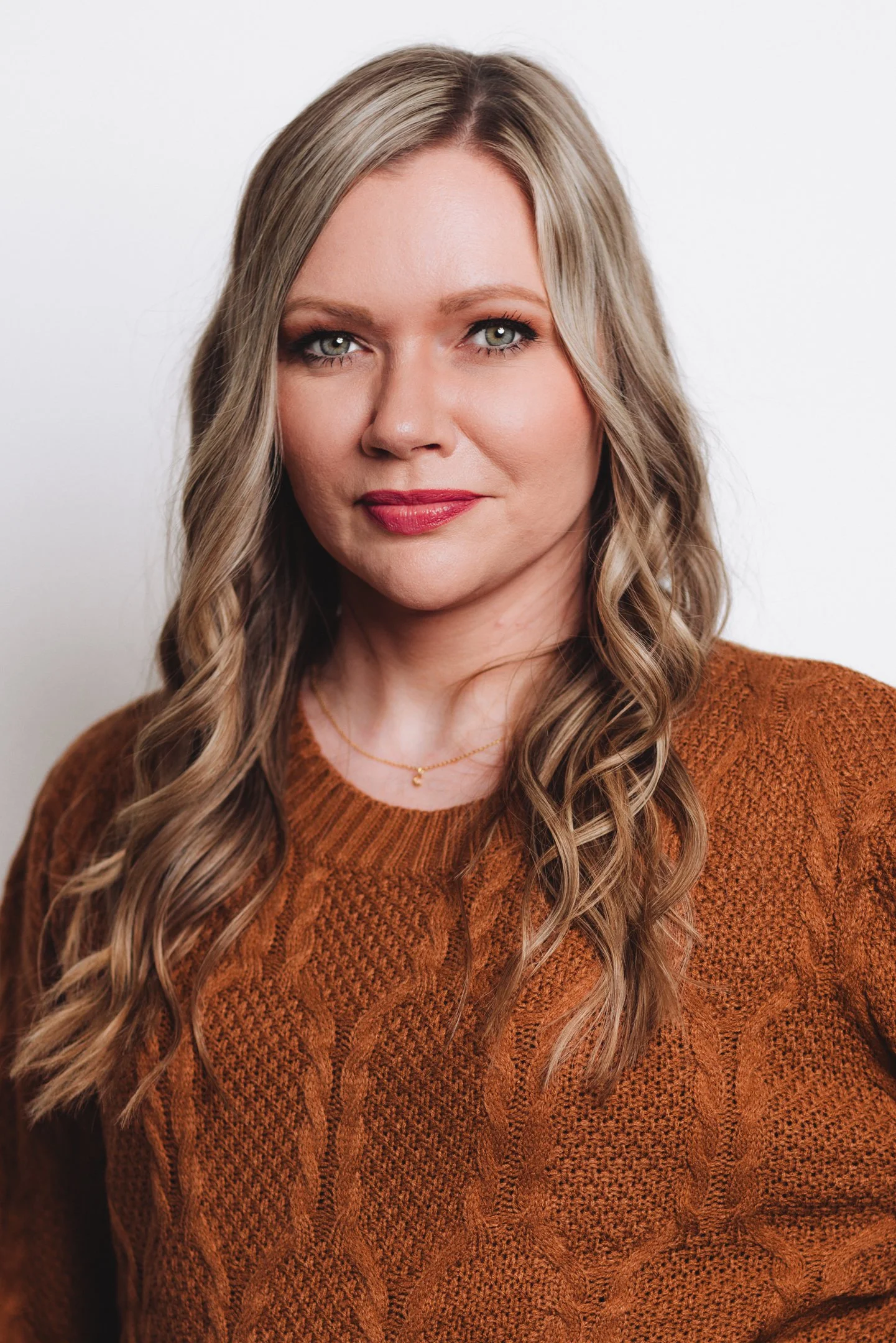 A woman with long, wavy blonde hair, wearing a rust-colored cable knit sweater and a delicate gold necklace, looking at the camera with a neutral expression against a plain white background.