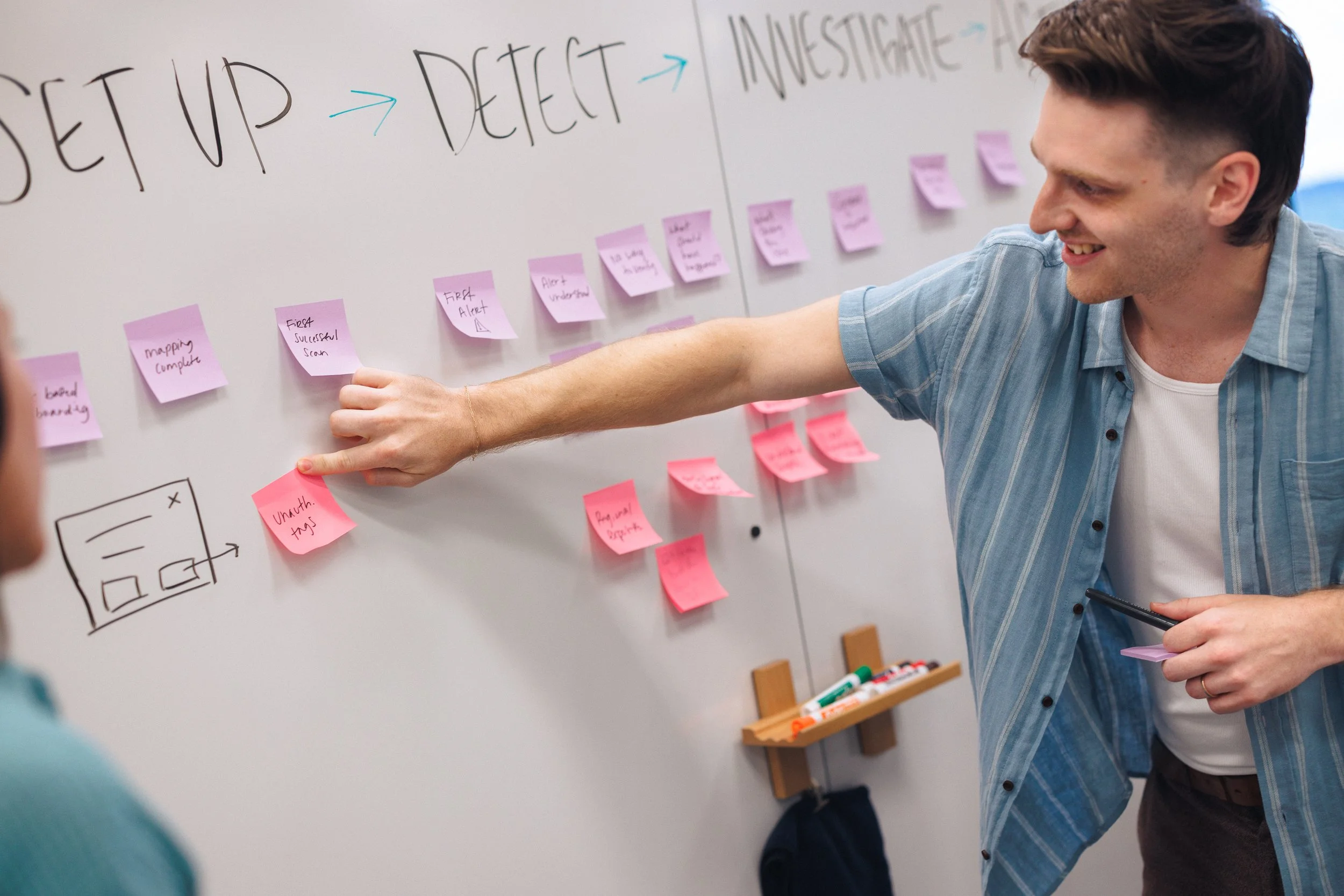 Man in a blue striped shirt smiling and pointing at pink sticky notes on a whiteboard with a flowchart and purple sticky notes above. The whiteboard has handwritten words 'SET UP', 'DETECT', and 'INVESTIGATE' connected with arrows.