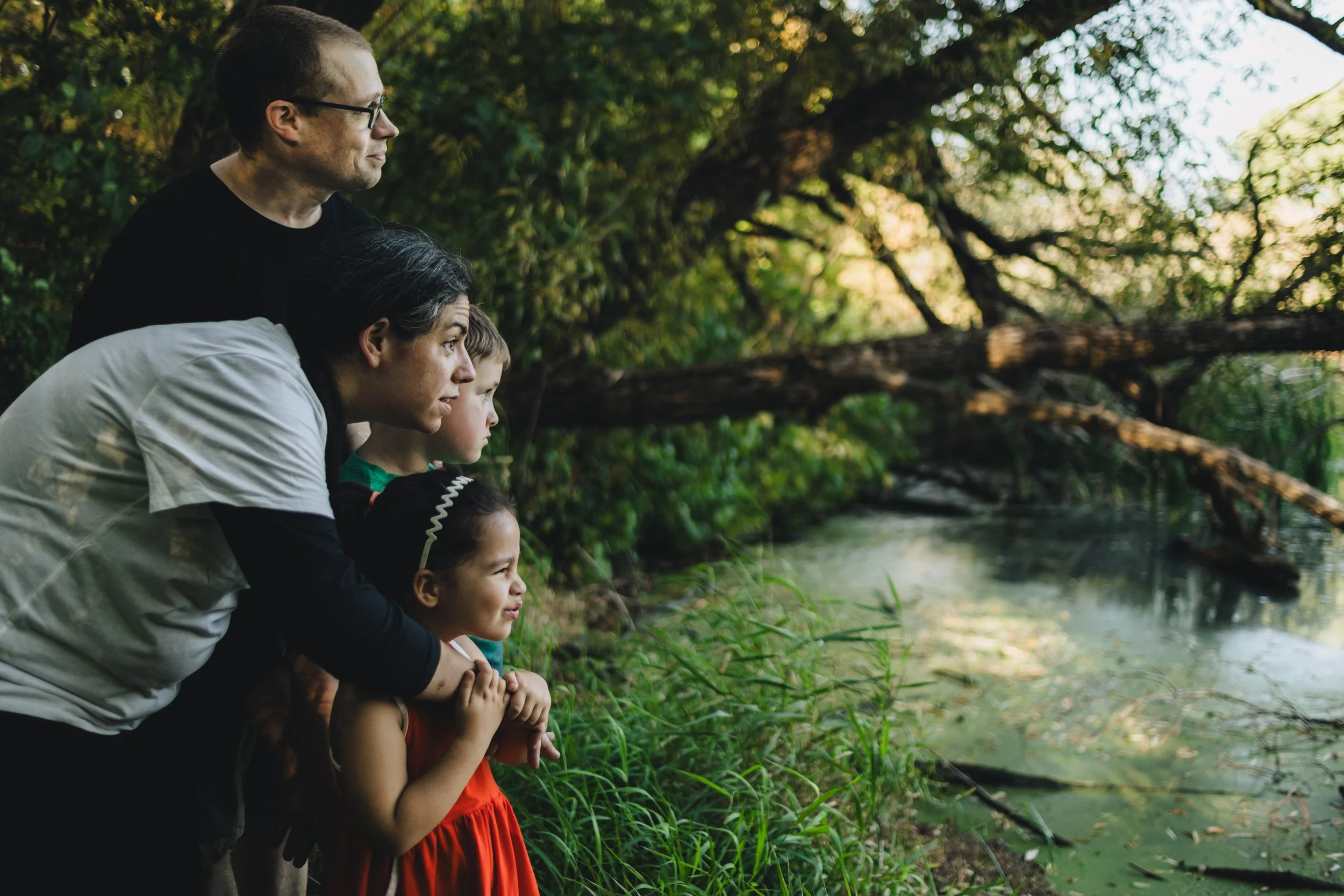 A family of five, including two adults and three children, stands by a riverbank, observing the water and trees.
