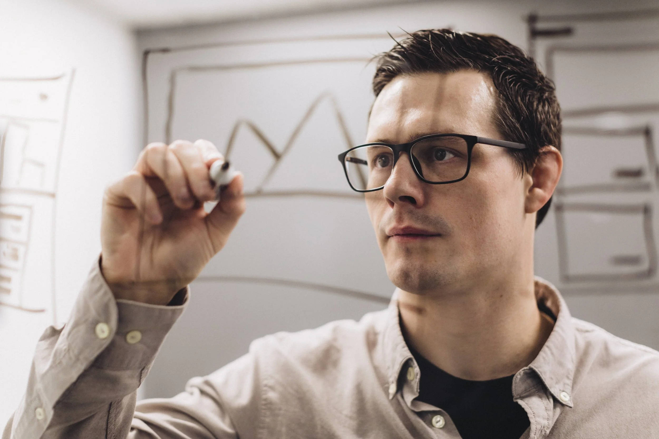 A man wearing glasses writes on a transparent surface with a marker, with charts and diagrams in the background.