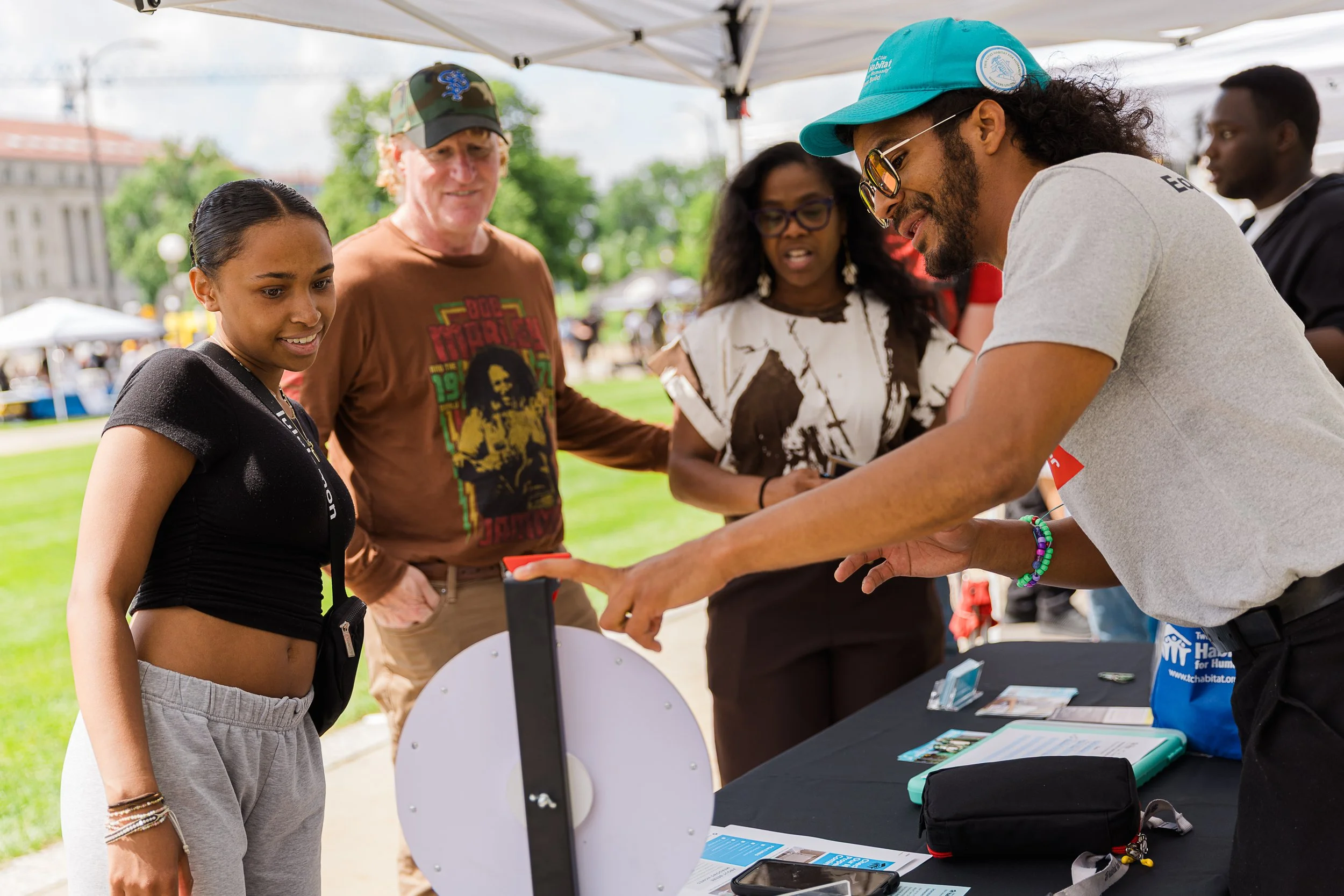 A group of people at an outdoor event under a tent, with a young woman listening to a man in sunglasses explaining something to her at a booth.