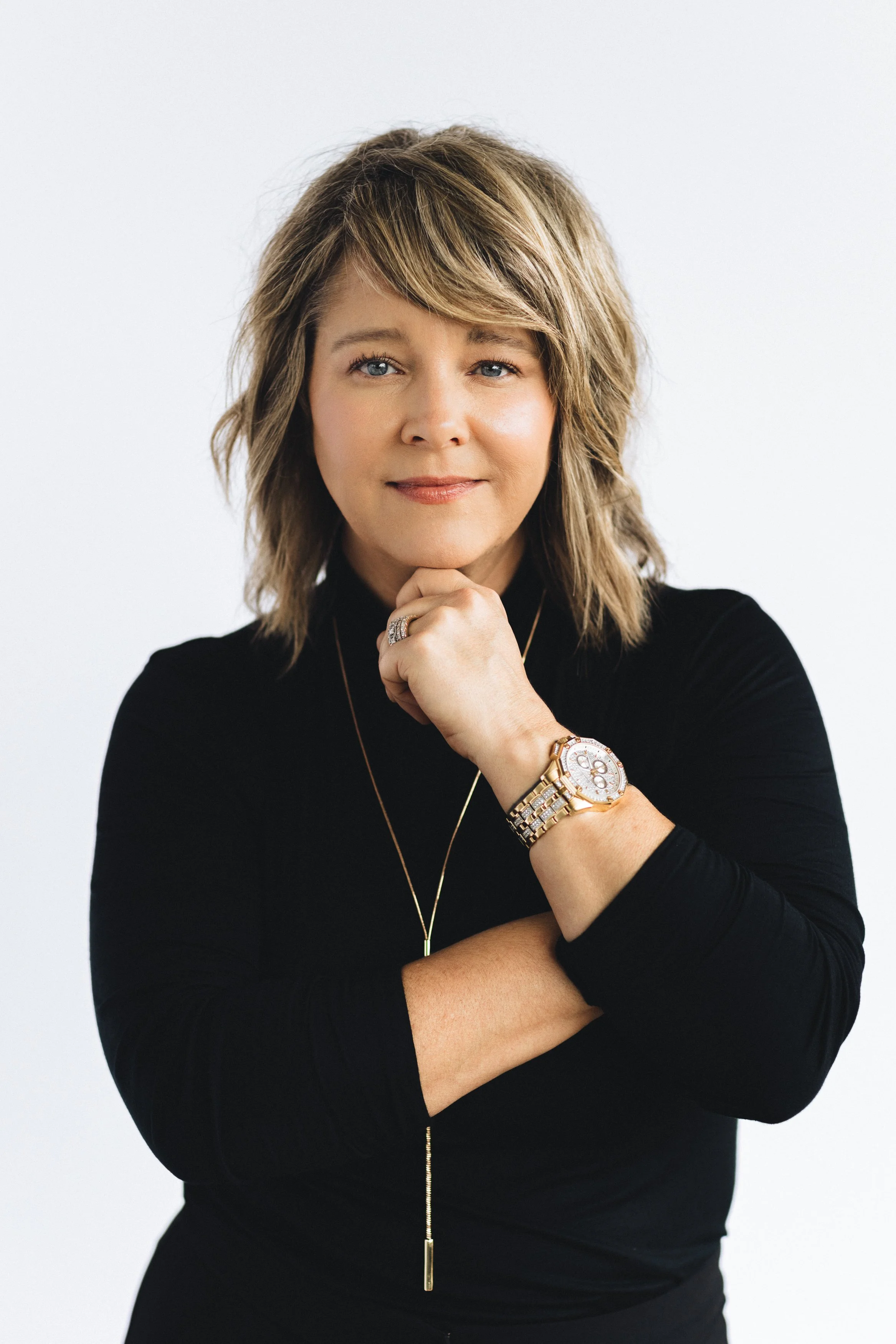 A woman with shoulder-length blonde hair and blue eyes, wearing a black top, multiple gold necklaces, a large gold watch, and a ring, standing against a white background. She is resting her chin on her right hand, with her left arm crossed, looking d