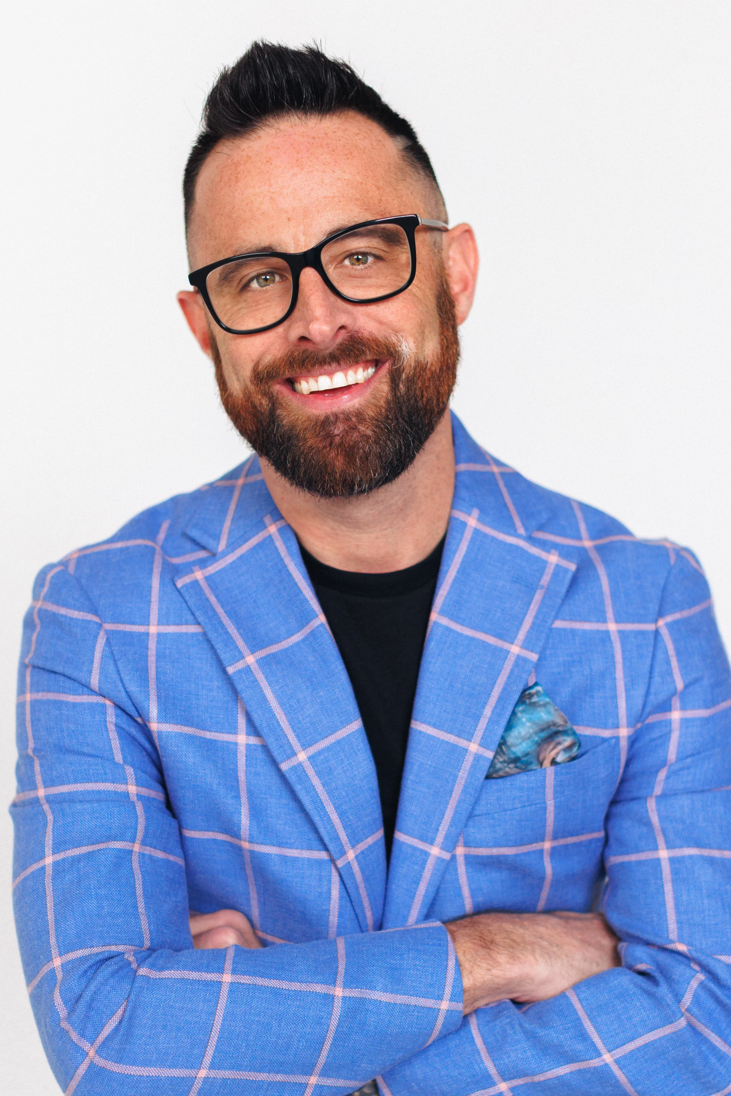 A smiling man with glasses, a beard, and short dark hair, wearing a blue checkered blazer and a black shirt, with arms crossed, posing against a white background.