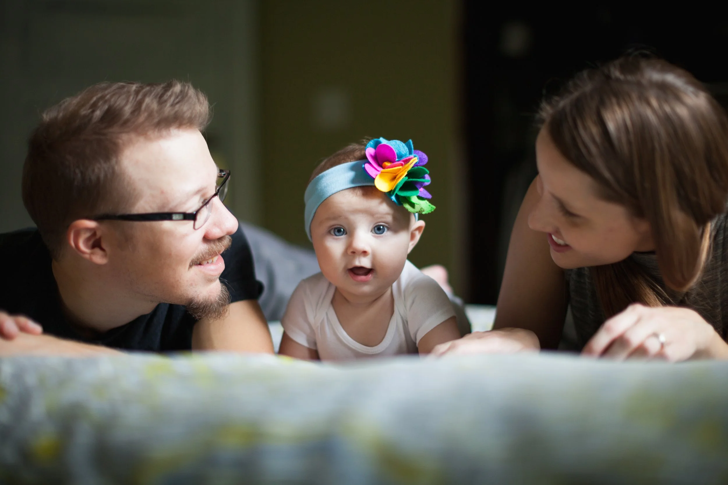 A baby girl with blue eyes, wearing a colorful flower headband, lying on her stomach between her smiling mother and father, who are looking at her affectionately.