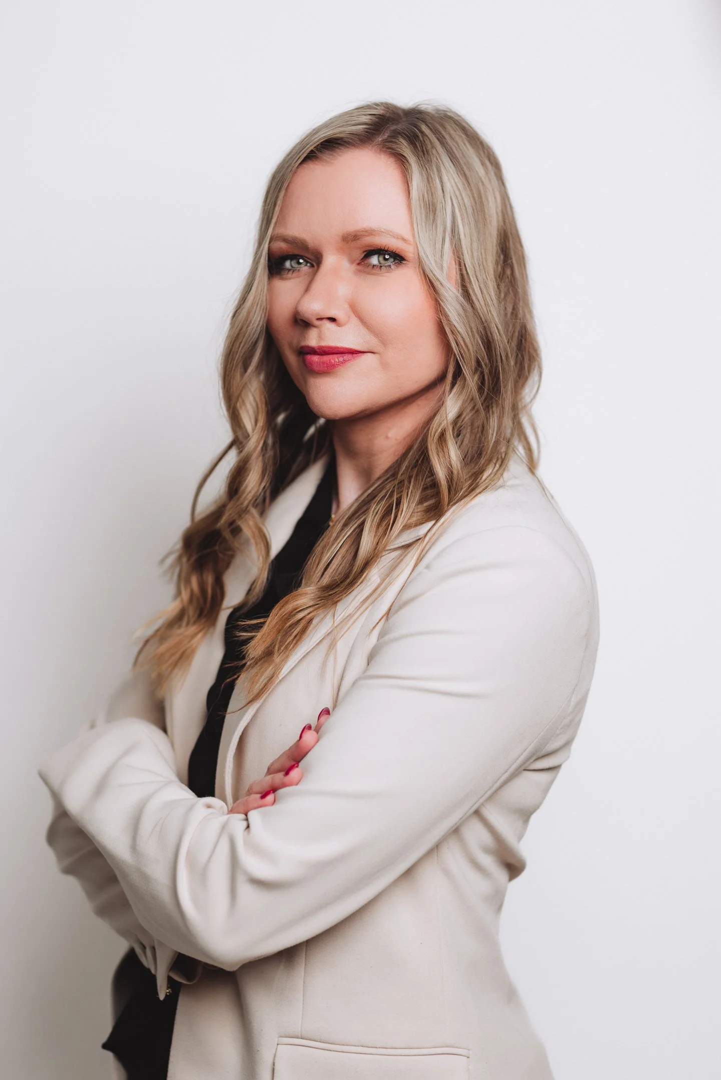 Portrait of a woman with blonde hair, crossed arms, wearing a cream blazer, standing against a white background.