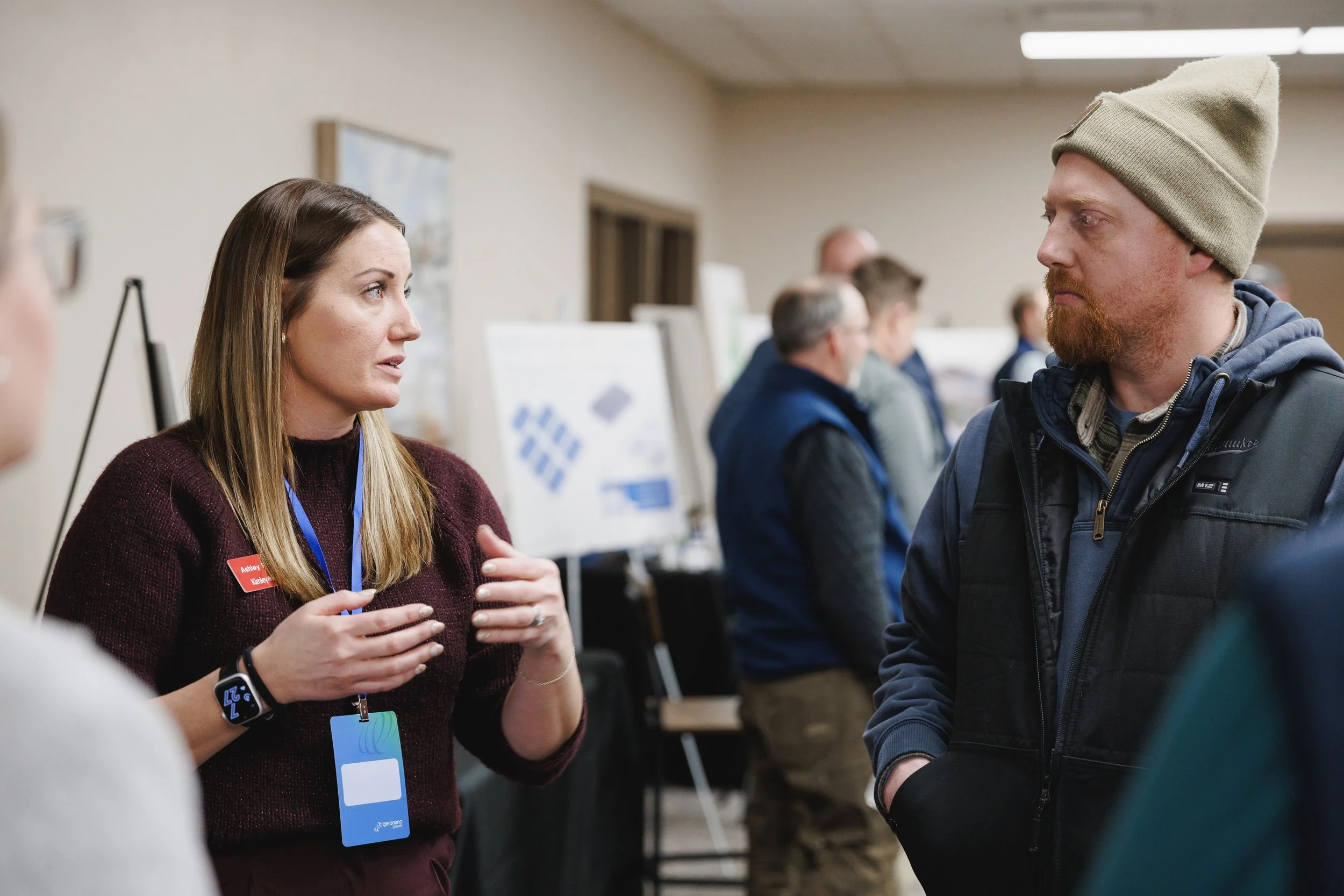 A woman with long brown hair wearing a burgundy sweater, speaking and gesturing with her hands to a man with a beard, light brown hair, wearing a beige beanie and a dark vest at an indoor event with posters and other people in the background.
