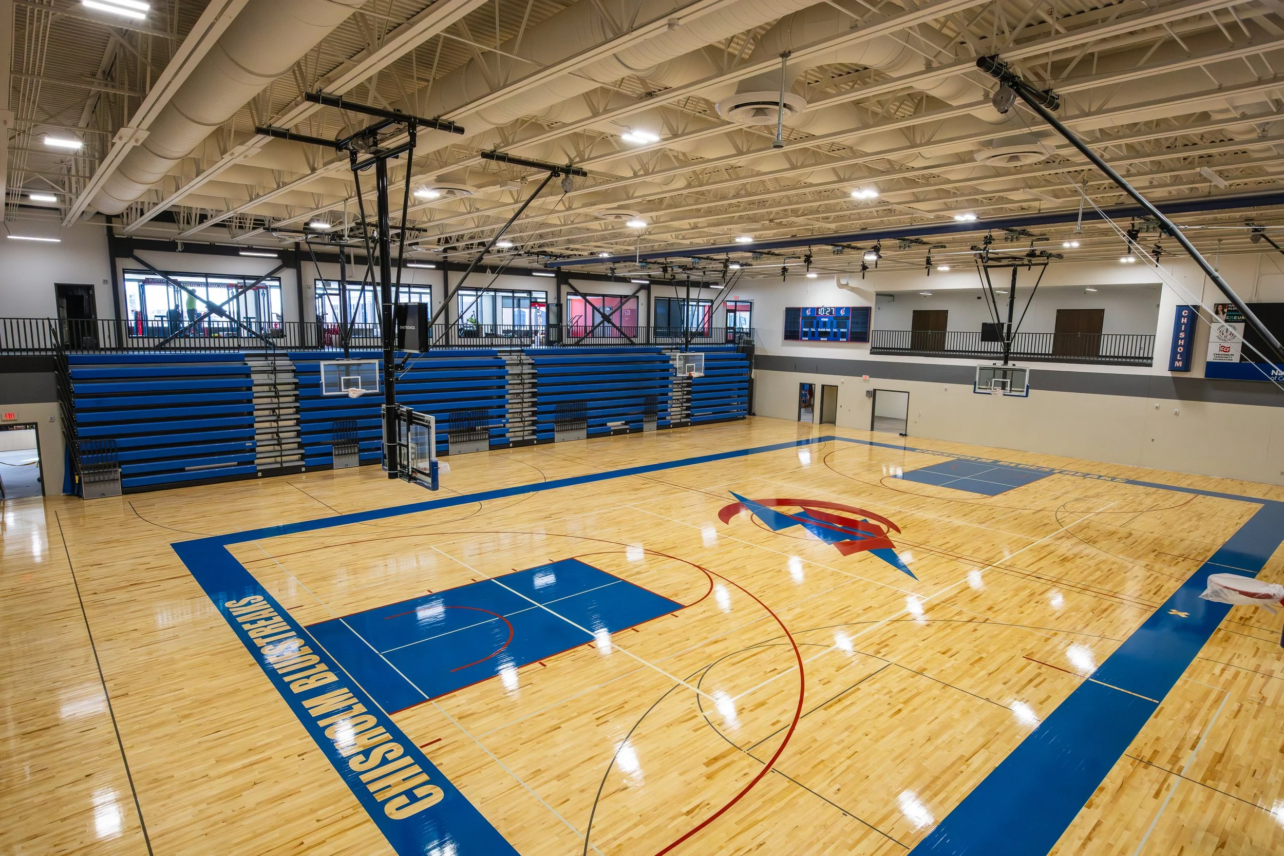 Empty indoor basketball court with blue and red markings, surrounded by blue bleachers and large windows, with a scoreboard on the wall.