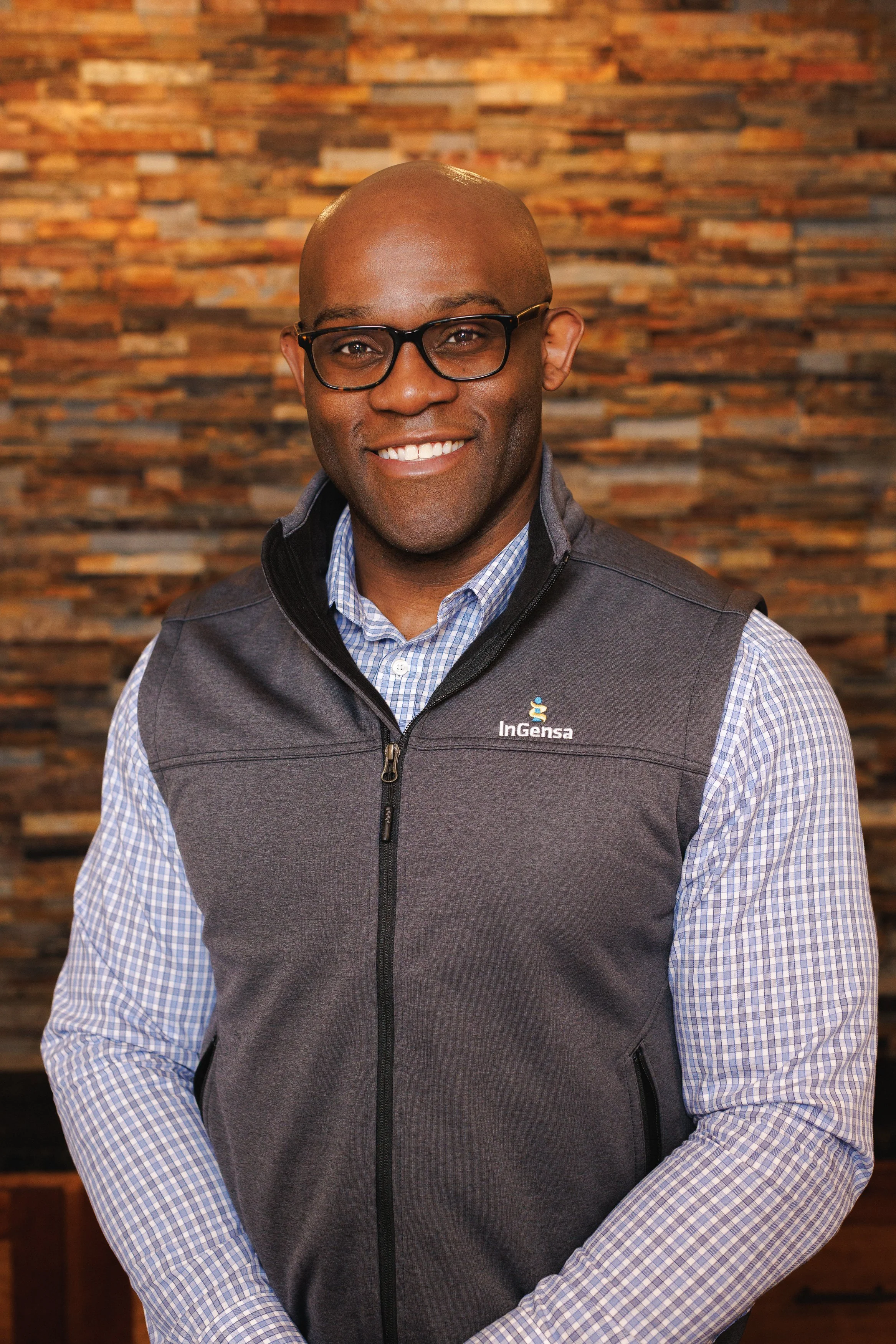 A smiling man wearing glasses, a gray vest with a logo that reads InGensa, and a light blue checkered shirt, standing in front of a textured brick wall background.