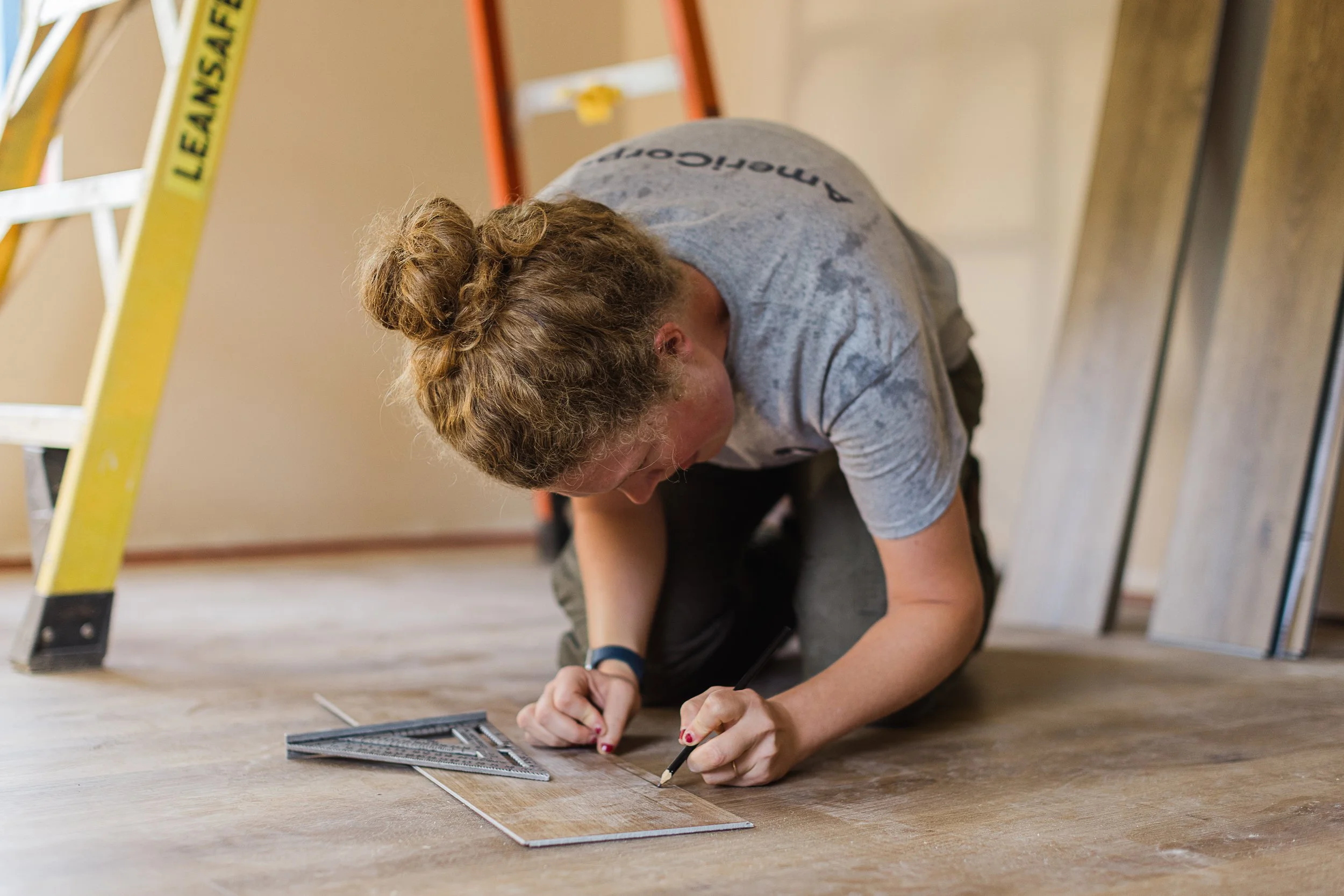 A woman with curly hair working on a flooring project on a wooden floor, using a square ruler and a pencil with a ladder nearby.
