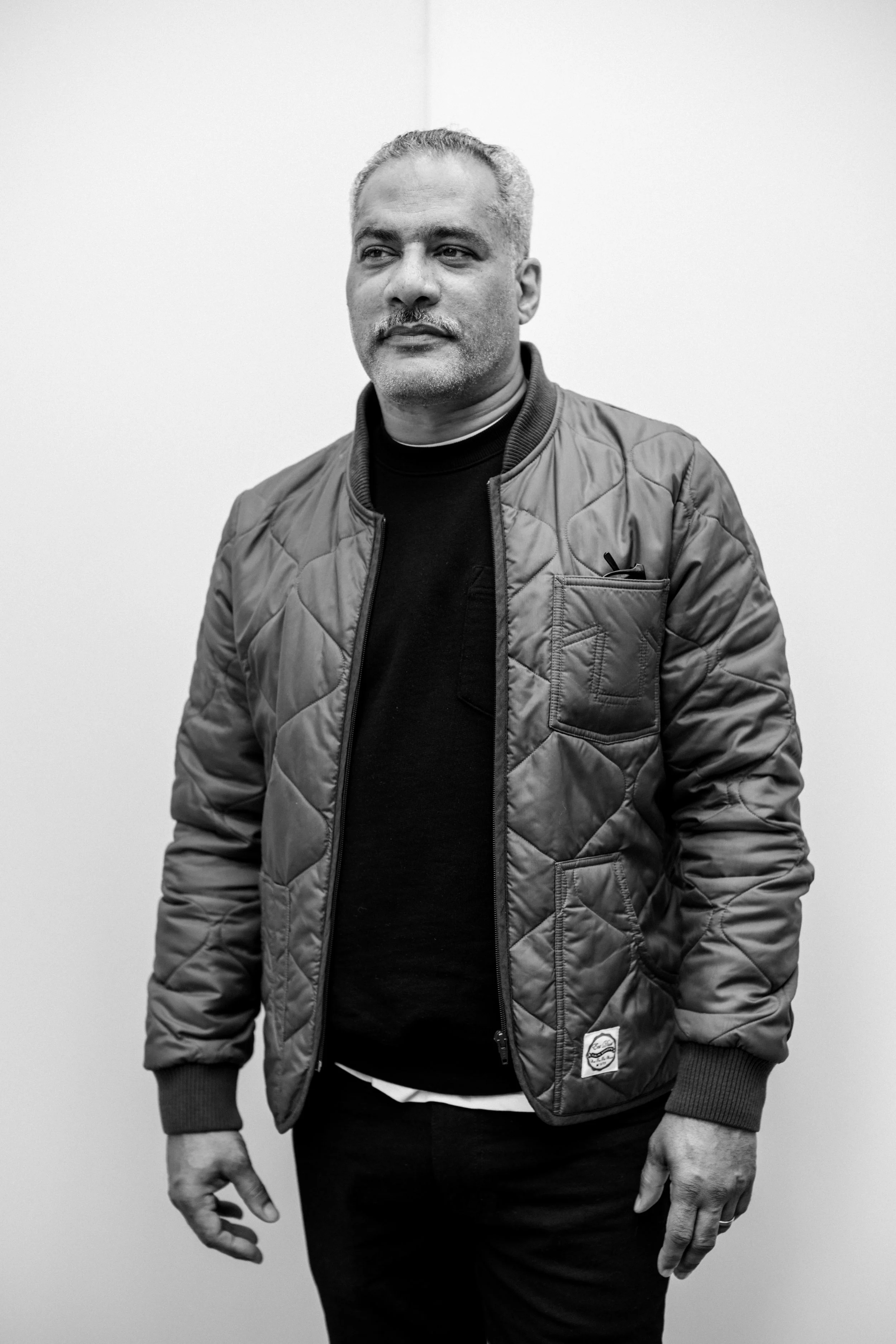 Black and white photo of a middle-aged man with short hair and a mustache, wearing a quilted jacket and dark clothing, standing against a plain light-colored background.
