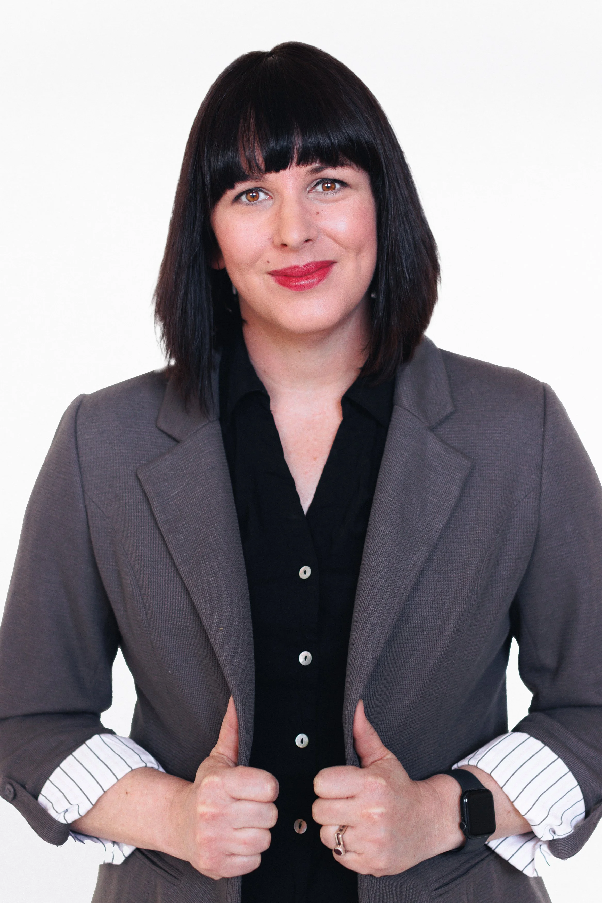 A woman with black hair and bangs, wearing a gray blazer over a black shirt, smiling, with her hands adjusting the blazer, standing against a plain white background.