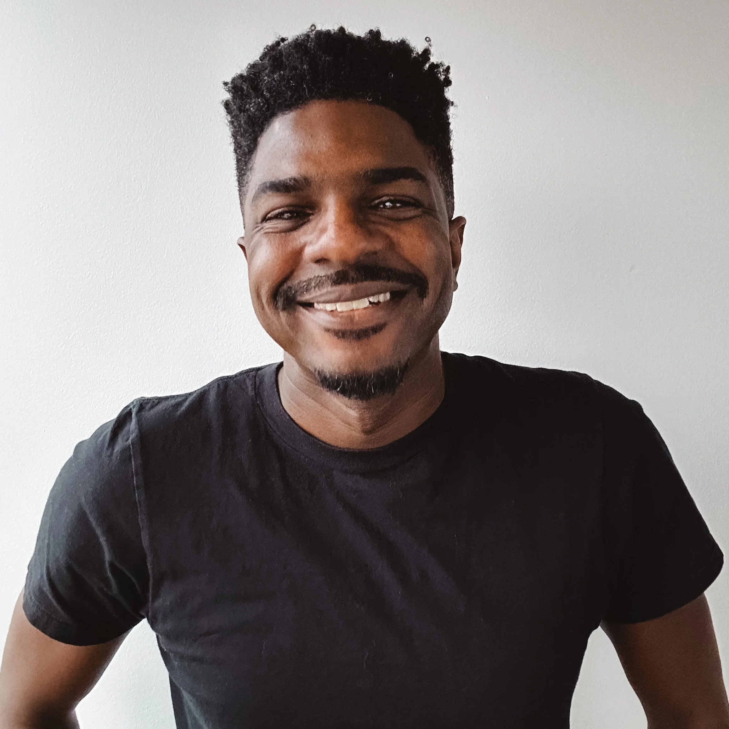 A young man with curly black hair, a goatee, and a mustache, smiling at the camera, wearing a black T-shirt, standing against a plain white wall.