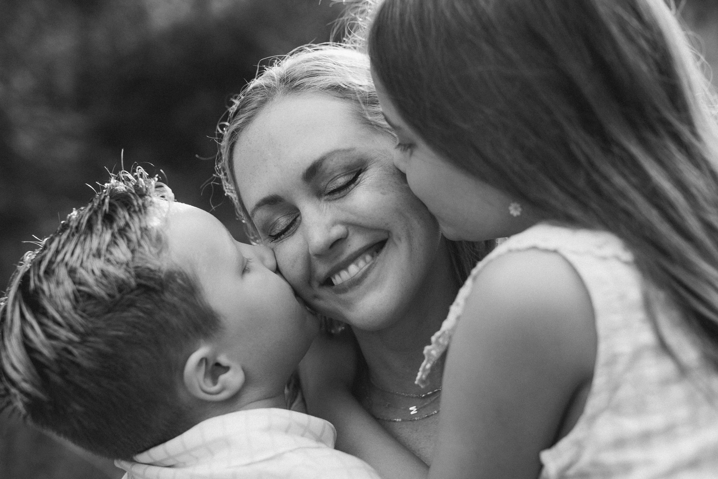 A woman being kissed on the cheek by two children, all smiling with eyes closed, in a close-up black and white portrait