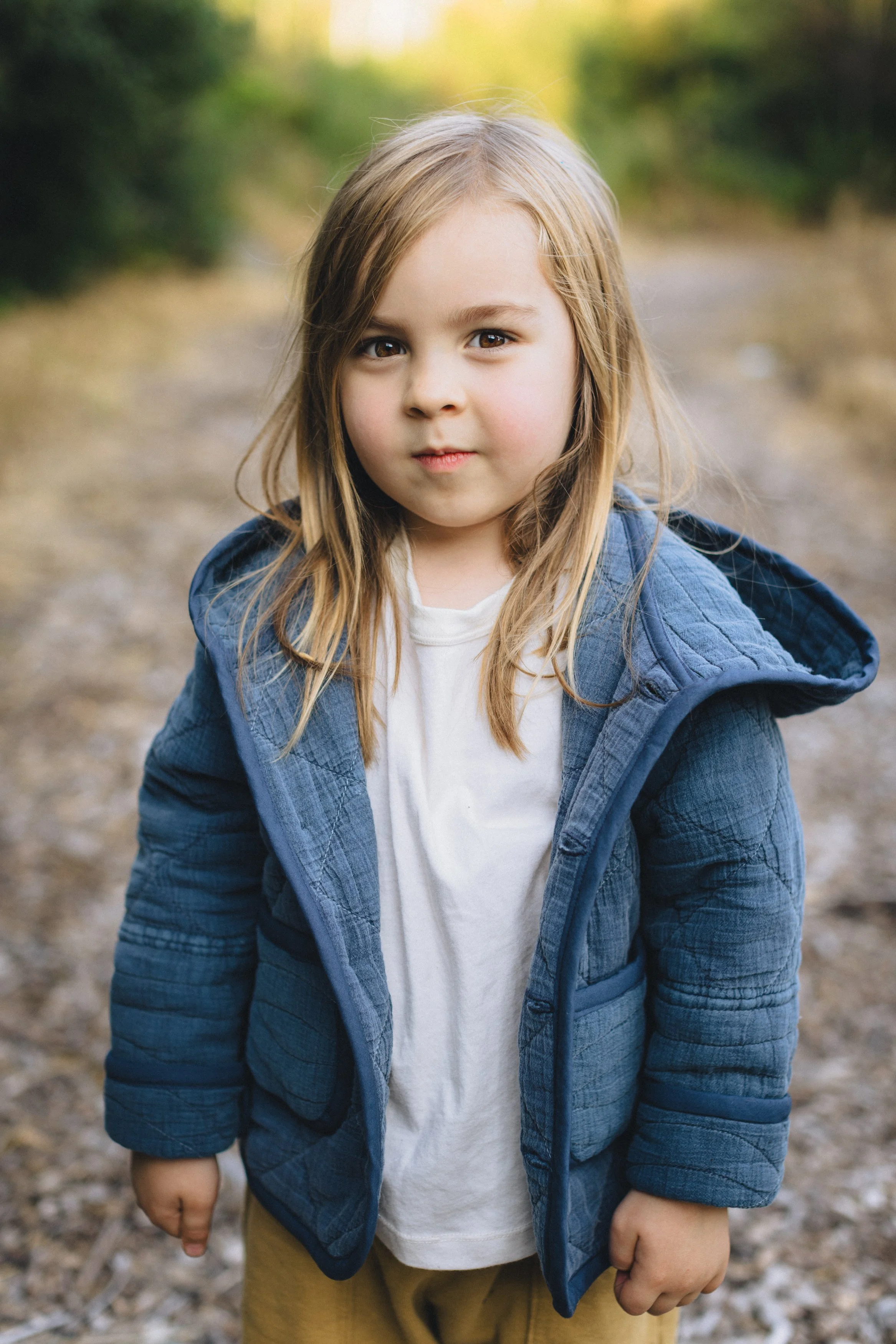 A young girl with long blonde hair wears a white shirt and a blue quilted jacket, standing outdoors on a dirt path with greenery in the background.
